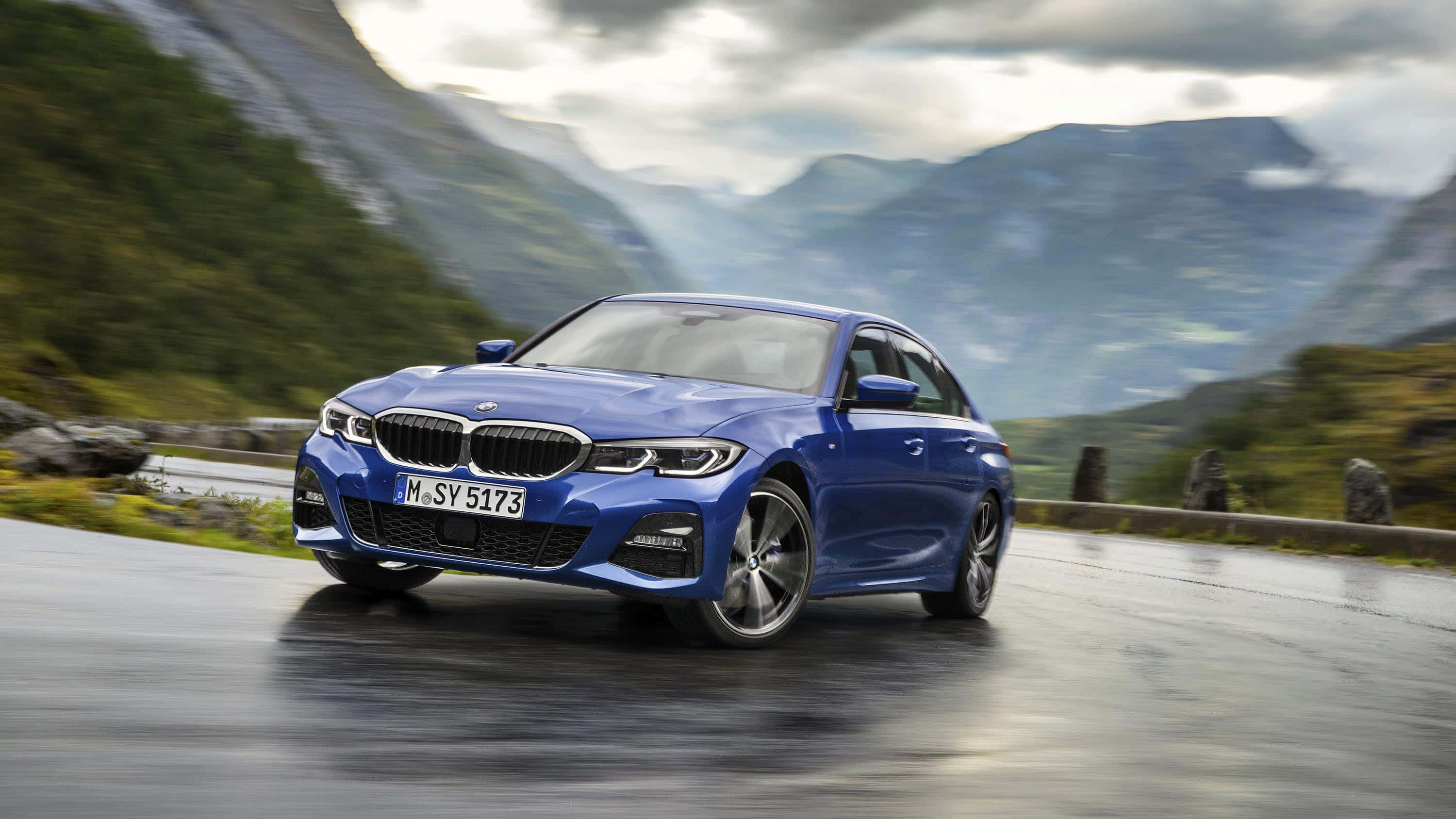 M Series 桌布 壁紙 — 一輛充滿活力的藍色BMW轎車在潮濕的山路上疾馳，兩旁是茂密的綠色山坡和霧氣繚繞的山峰，天空多雲。