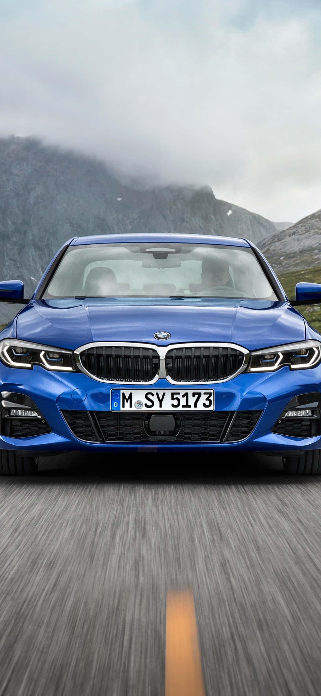 iPhone X Bmw 桌布 壁紙 — 一輛充滿活力的藍色BMW轎車在霧氣繚繞的山峰背景下沿山路飛馳。