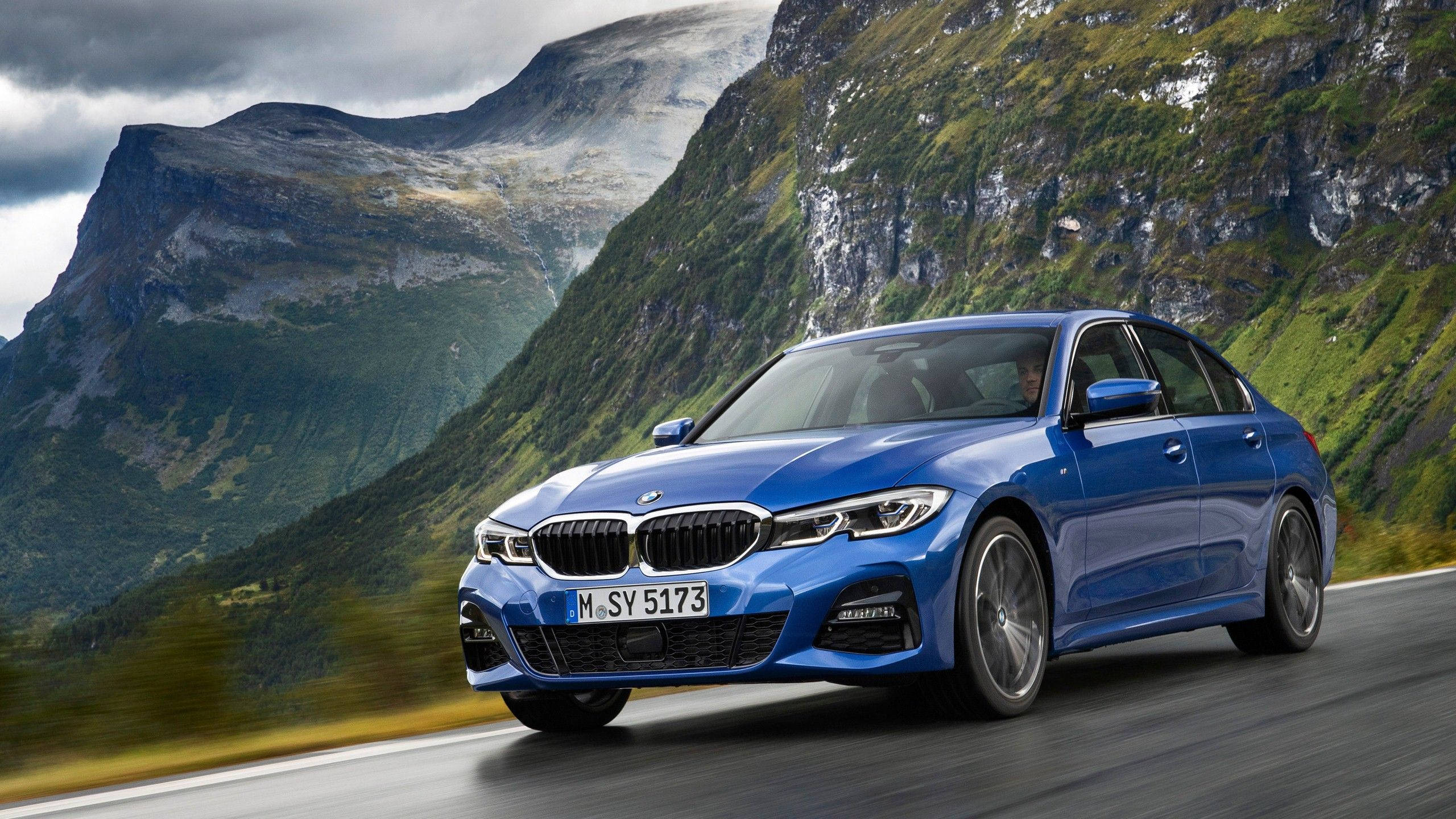 Bmw Cars 桌布 壁紙 — 一輛充滿活力的藍色BMW轎車沿著蜿蜒的山路飛馳，背景是鬱鬱蔥蔥的綠色懸崖和多雲的天空。