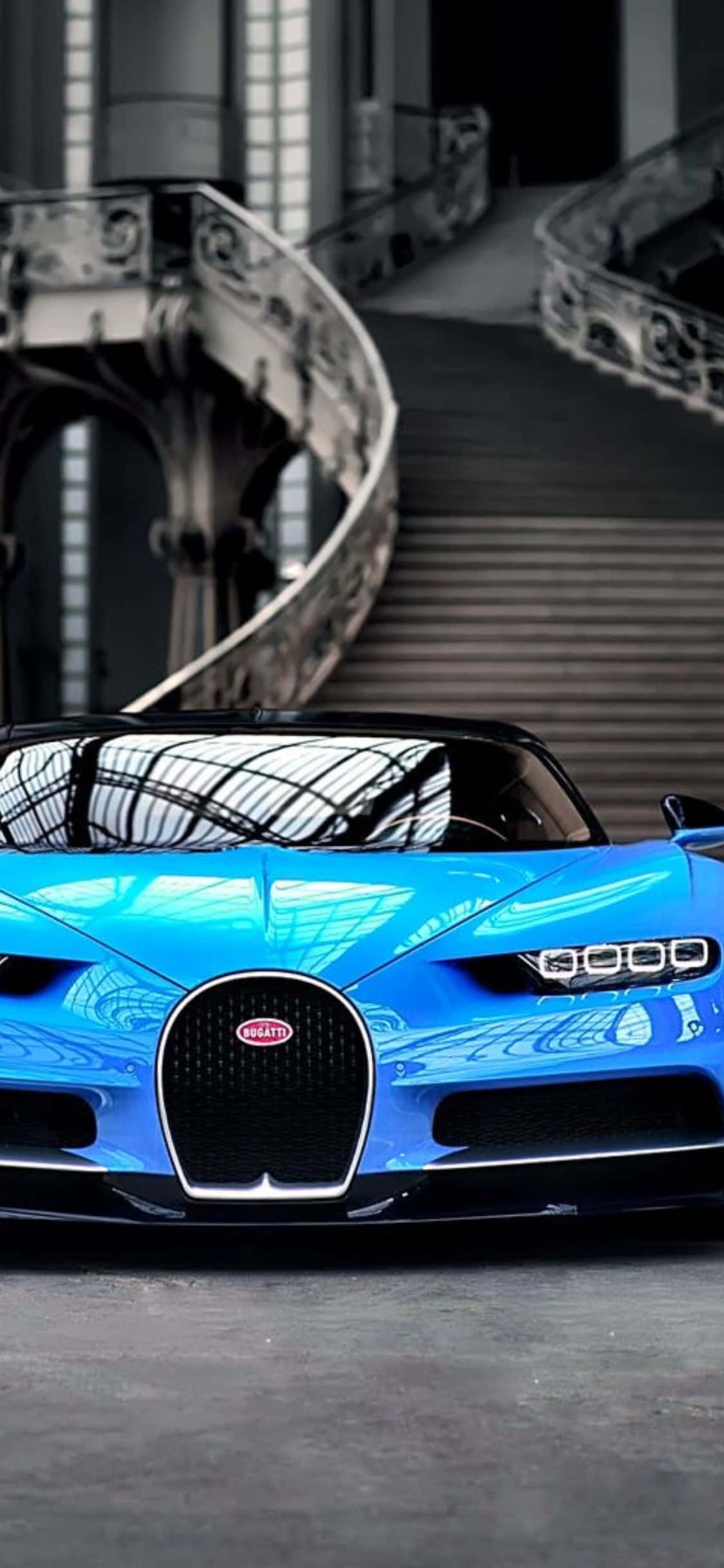 iPhone Xs Bugatti 桌布 壁紙 — 一輛充滿活力的藍色Bugatti跑車在明亮的燈光下，在一個擁有華麗螺旋樓梯的宏偉建築空間中閃閃發光。