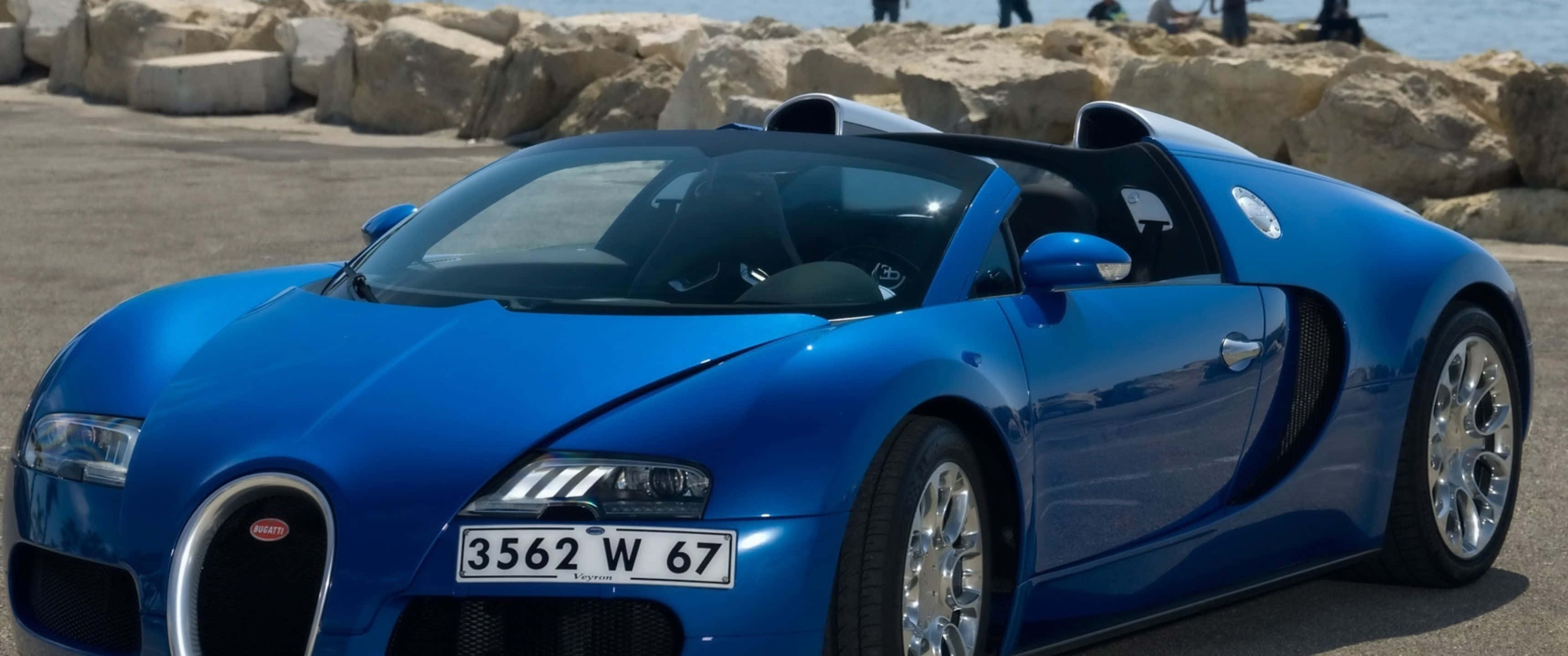 3440x1440p Bugatti 桌布 壁紙 — 一輛充滿活力的藍色Bugatti Veyron超級跑車在明亮的陽光下閃閃發光，停泊在岩石海邊懸崖附近。