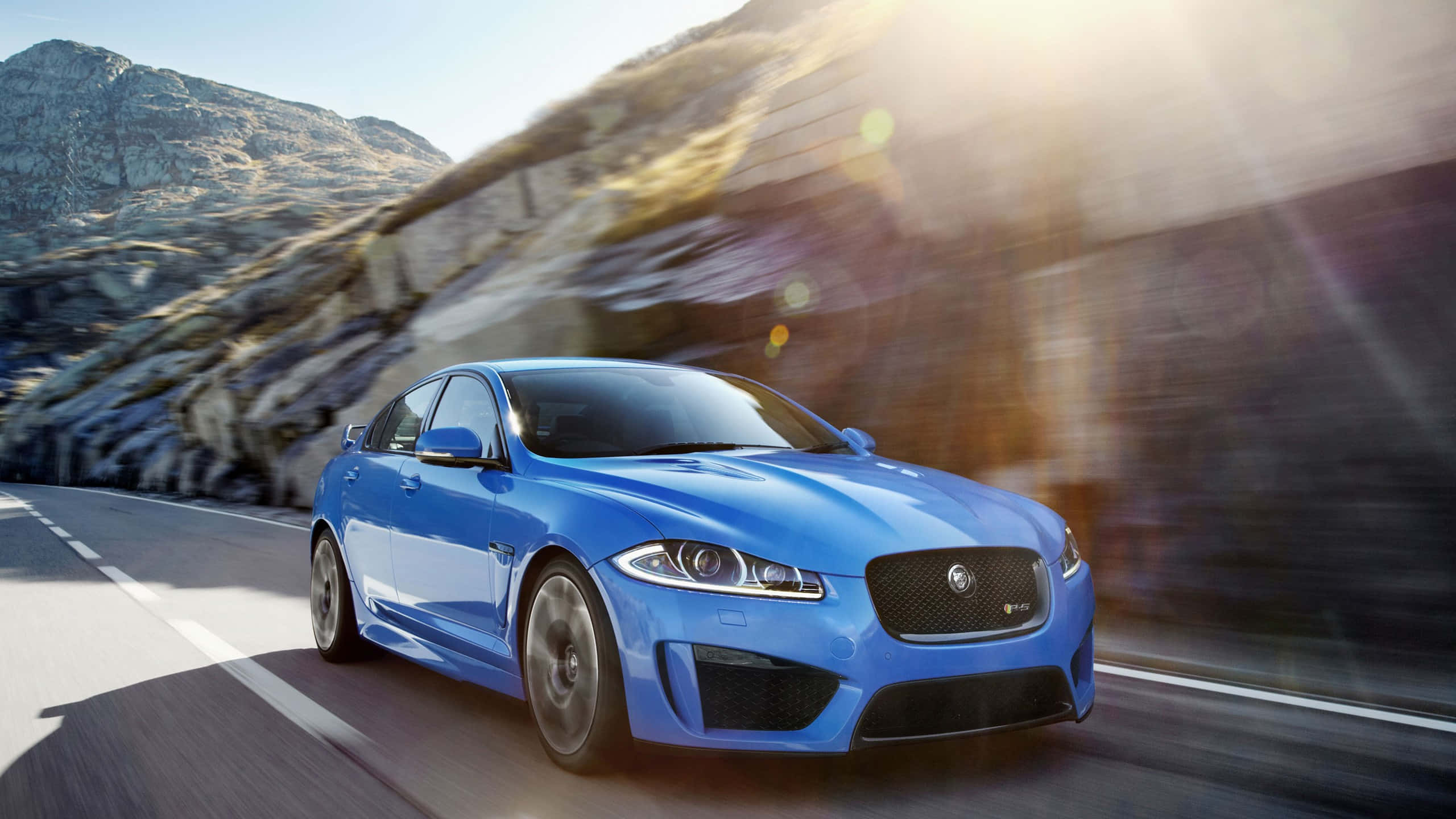 Jaguar Xfr 桌布 壁紙 — 一輛充滿活力的藍色Jaguar XF Sportbrake沿著山路疾馳，伴隨著太陽耀斑和動態模糊效果。
