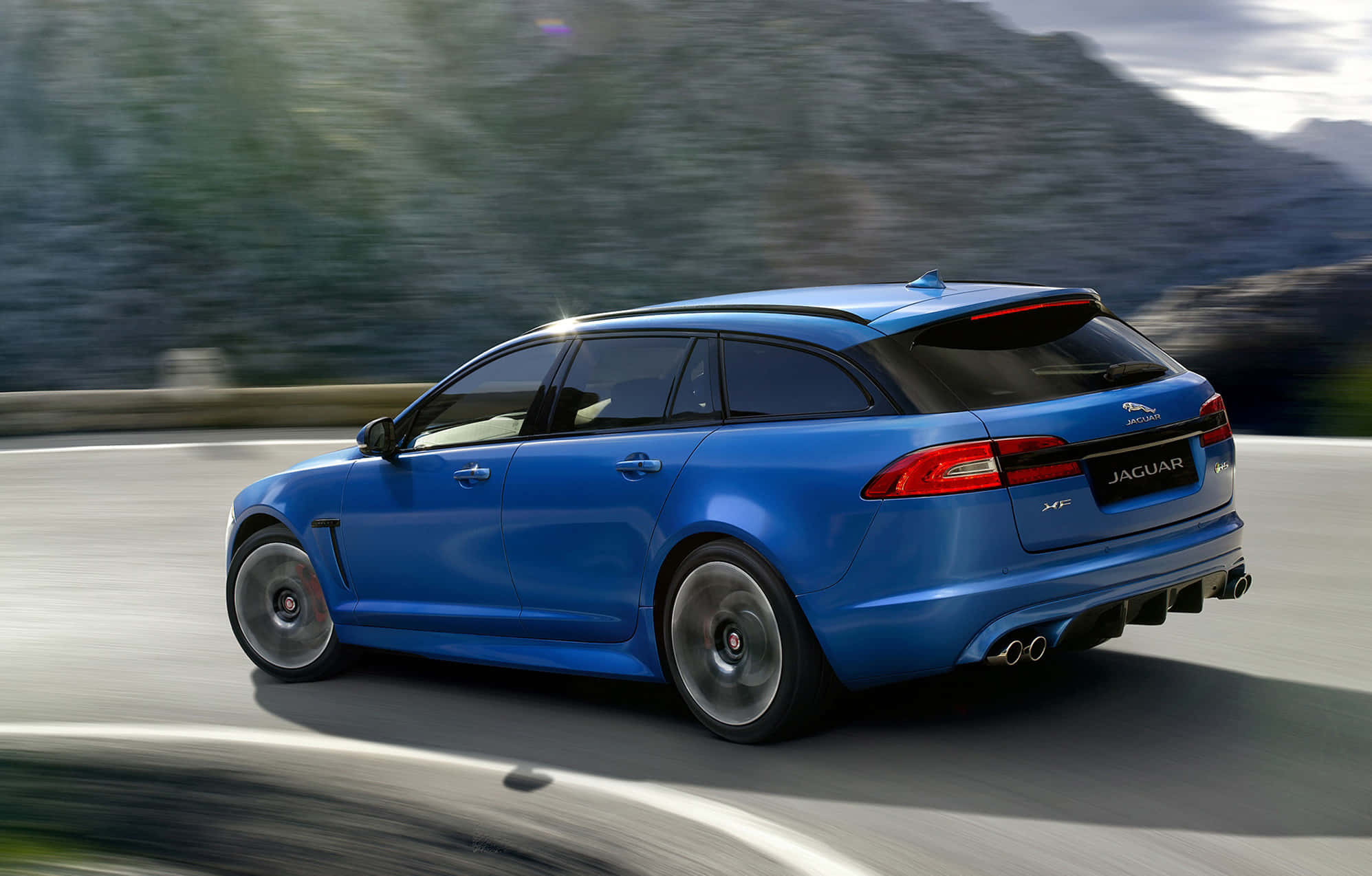 Jaguar Xfr 桌布 壁紙 — 一輛充滿活力的藍色Jaguar XF Sportbrake在蜿蜒的山路上疾馳，背景是模糊的綠色植物。