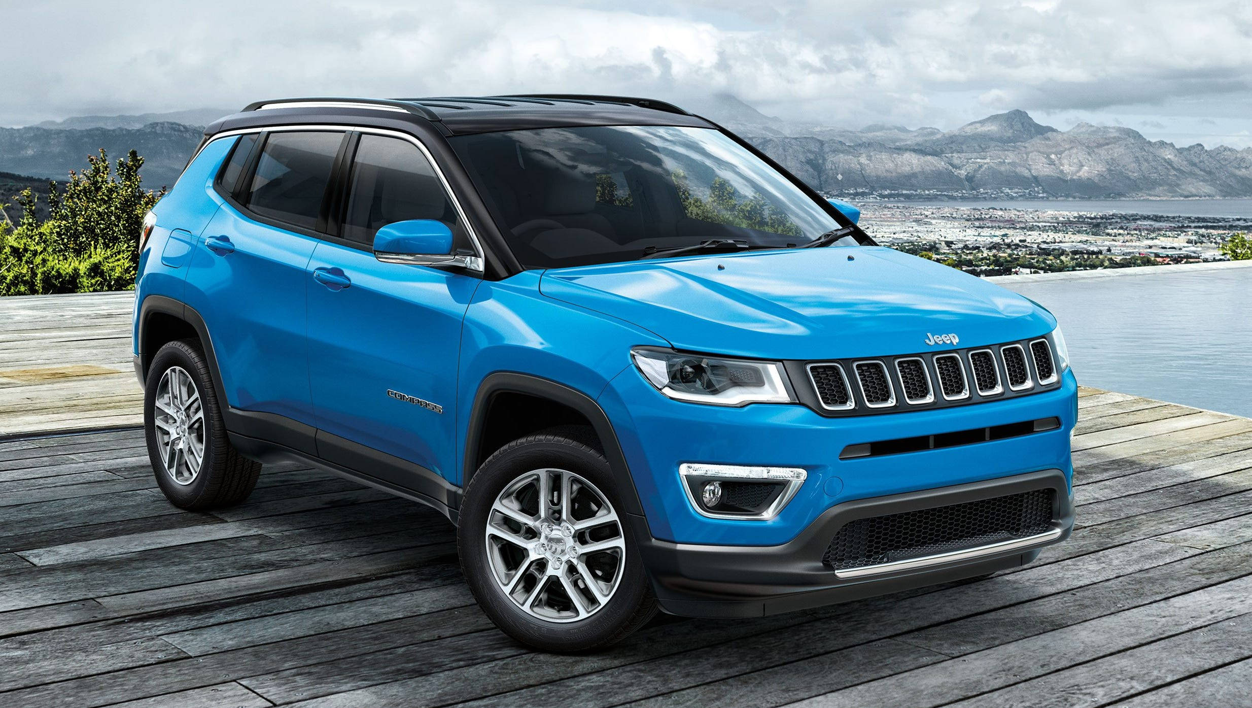 木紋 桌布 壁紙 — 一輛充滿活力的藍色Jeep Compass SUV停泊在木甲板上，俯瞰雲層下的沿海城市和山脈。