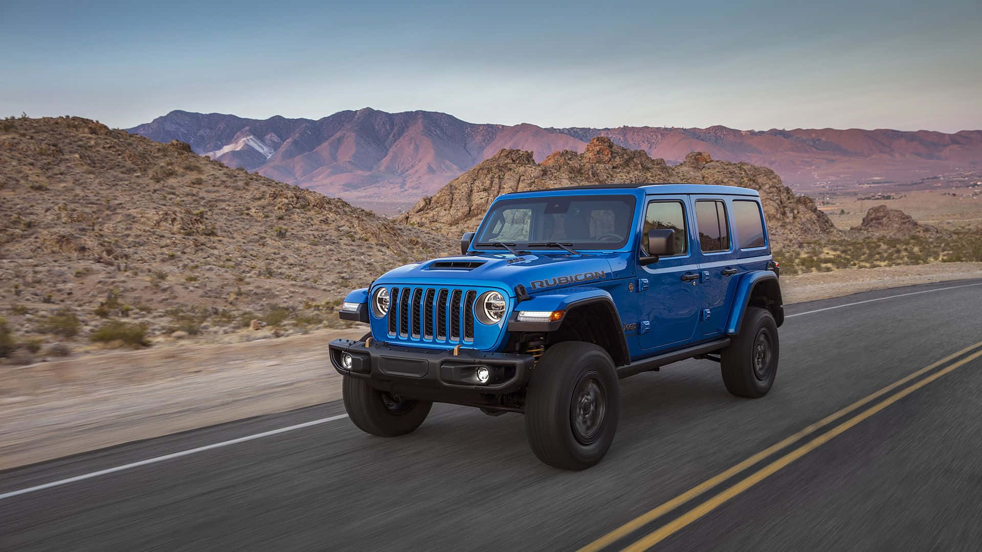 吉普 桌布 壁紙 — 一輛充滿活力的藍色Jeep Wrangler在沙漠公路上飛馳，背景是崎嶇的山脈和日落。