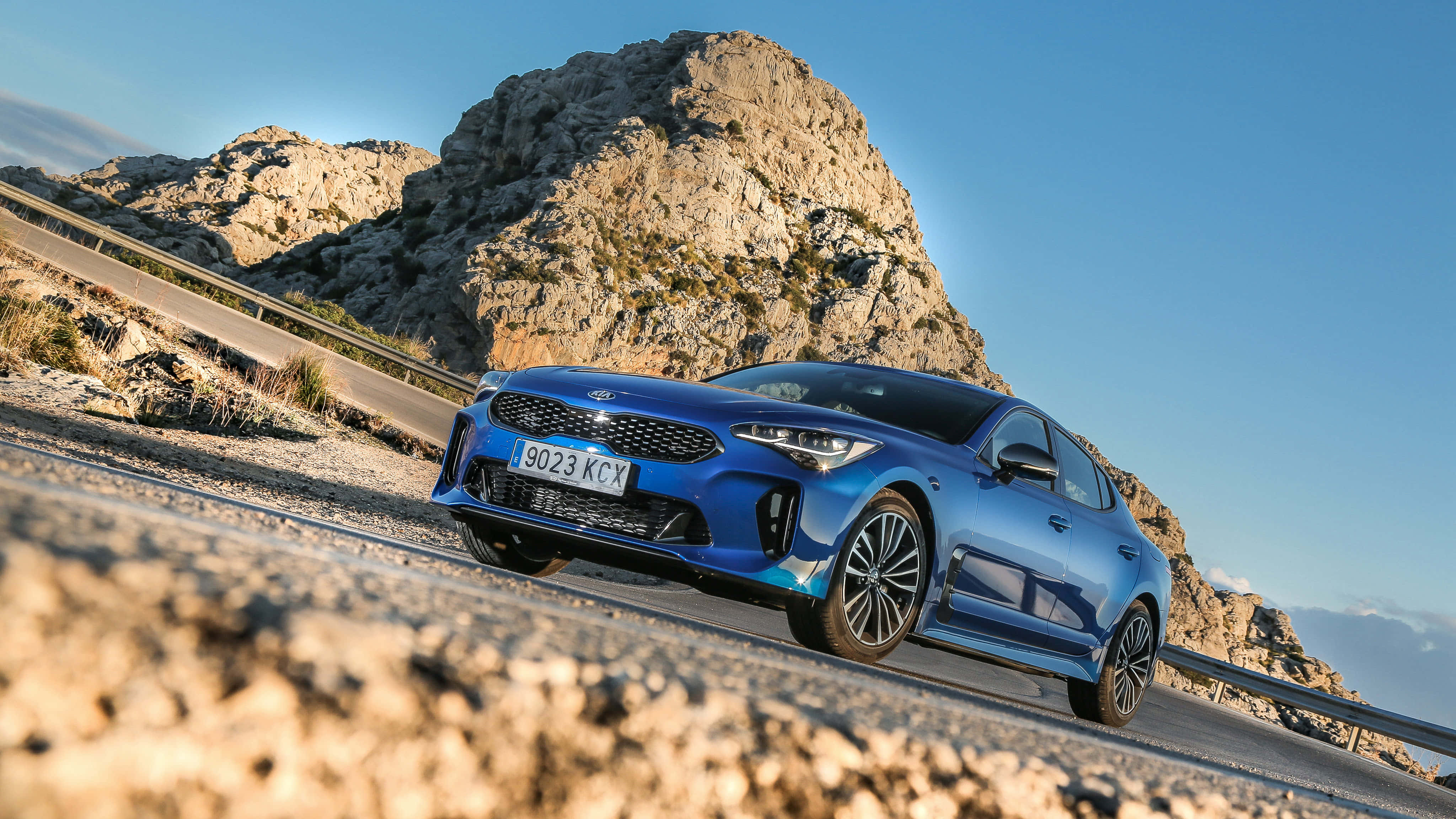 Kia Stinger 桌布 壁紙 — 一輛充滿活力的藍色Kia轎車在晴朗的藍天下沿著蜿蜒的山路行駛，背景是崎嶇的岩石地貌。
