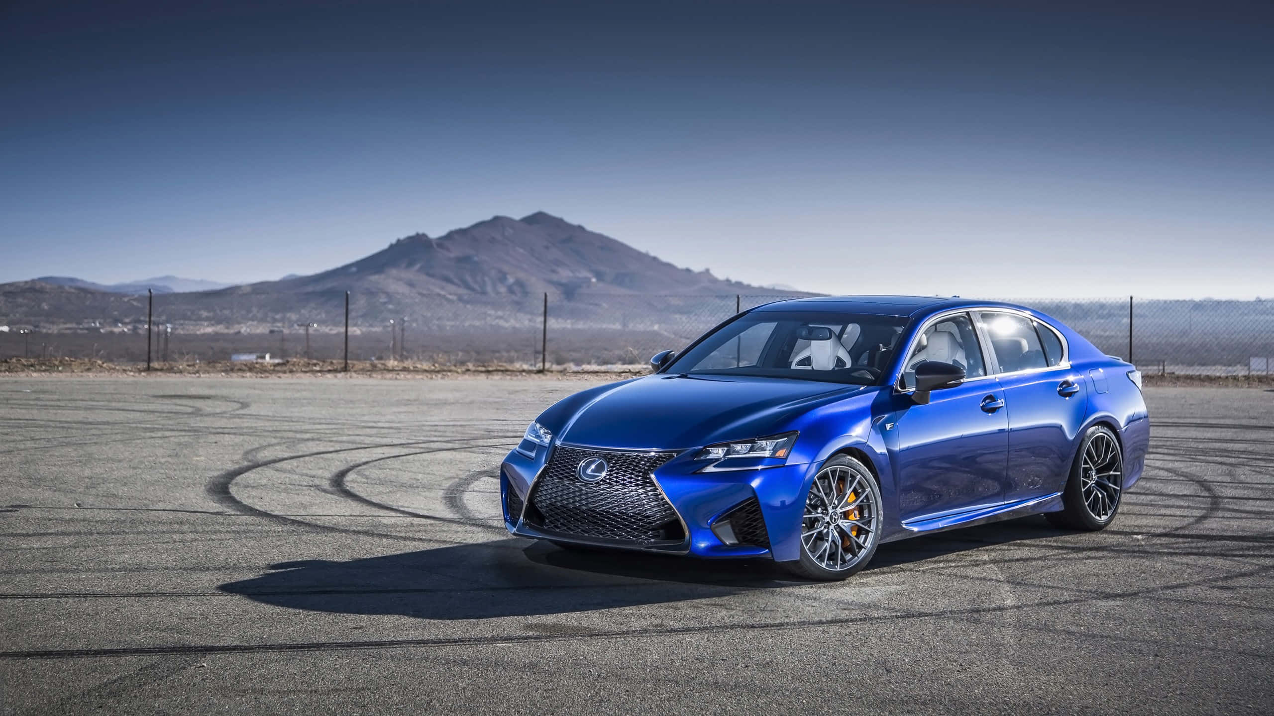 Lexus Gs 桌布 壁紙 — 一輛充滿活力的藍色Lexus RC F跑車在晴朗天空下，於鋪設的賽道上進行飄移動作，背景是山區地形。