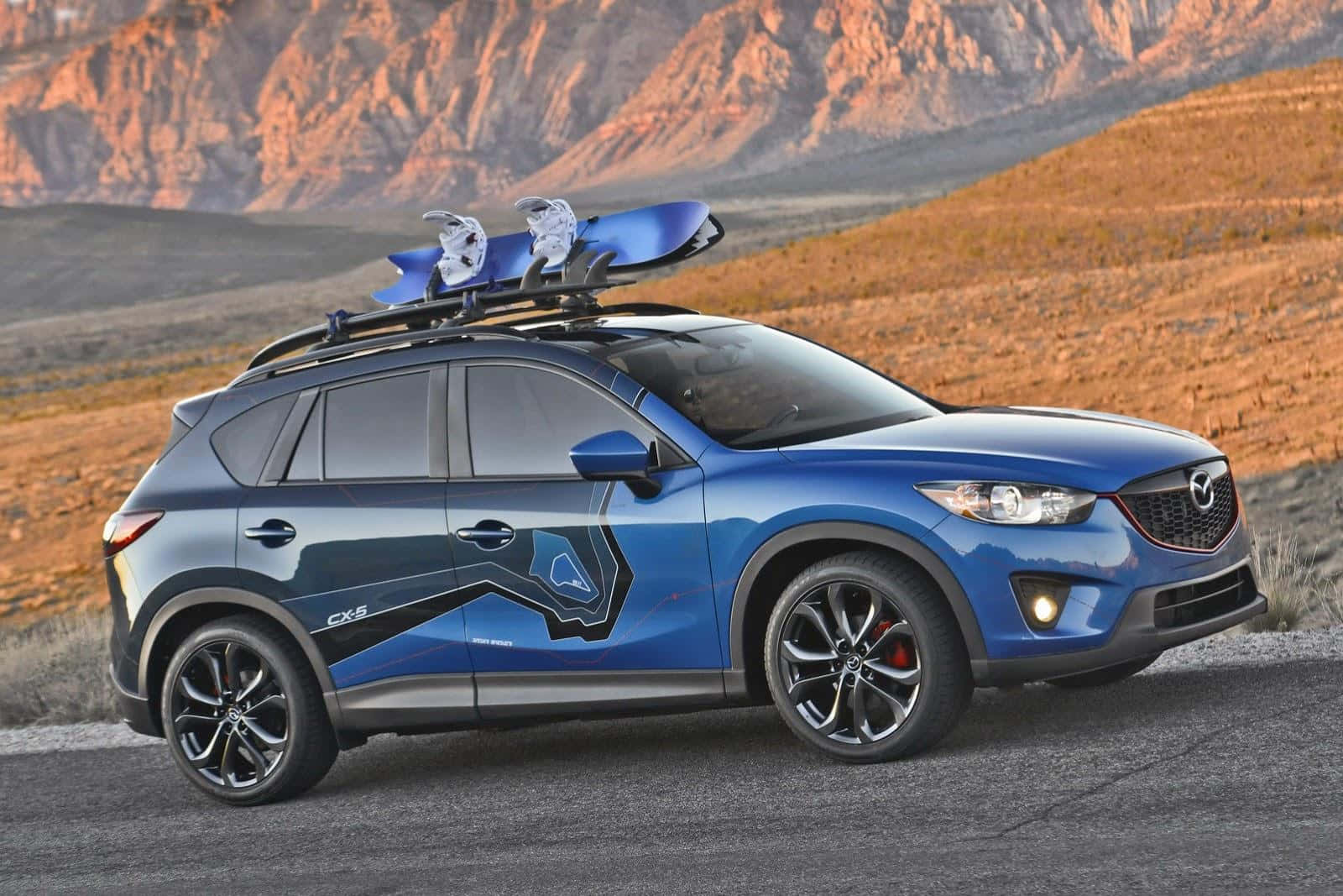Mazda Cx-5 桌布 壁紙 — 一輛充滿活力的藍色Mazda CX-5 SUV，車頂架上放著滑雪板，停在沙漠公路上，背景是崎嶇的橙色山脈，正值日落時分。