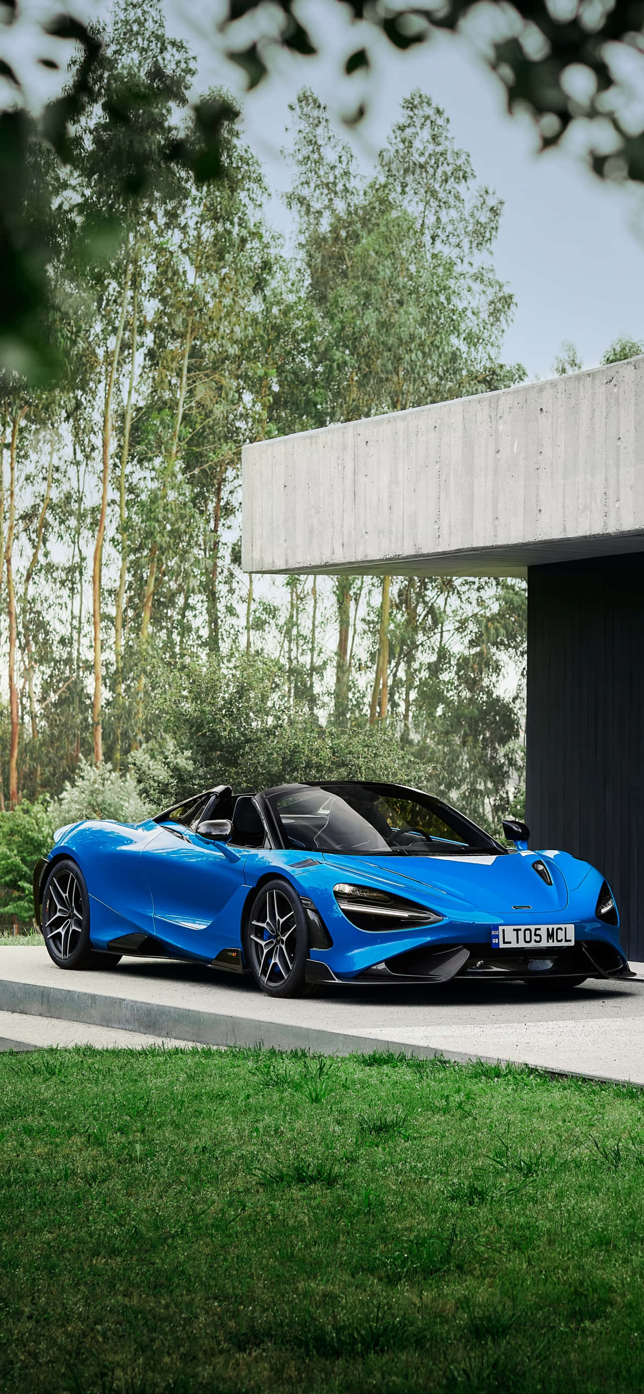 跑車 桌布 壁紙 — 一輛充滿活力的藍色McLaren 720S Spider停放在現代混凝土房屋旁的混凝土車道上，背景是茂密的綠色樹木。