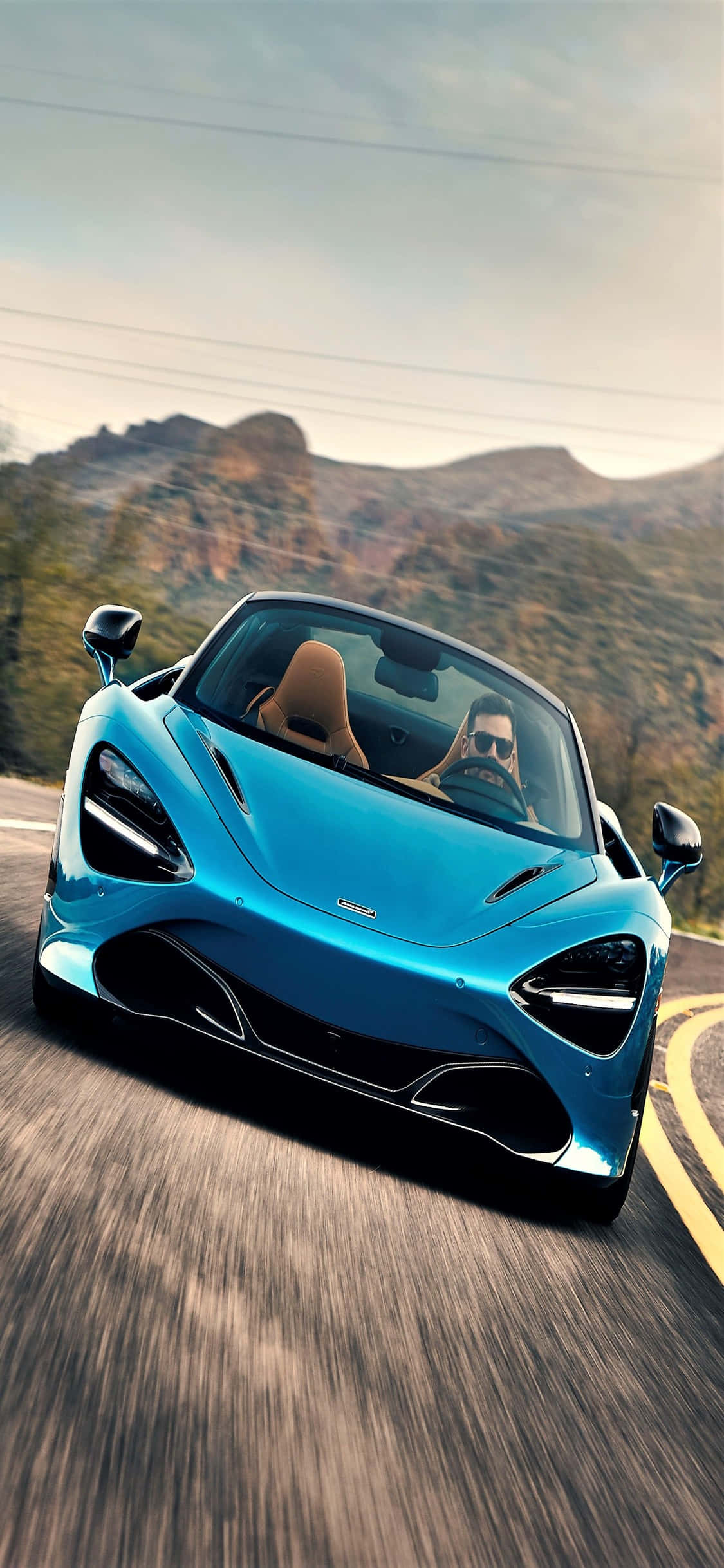 iPhone X Mclaren 720s 桌布 壁紙 — 一輛充滿活力的藍色McLaren跑車沿著蜿蜒的山路疾馳，司機戴著太陽眼鏡。