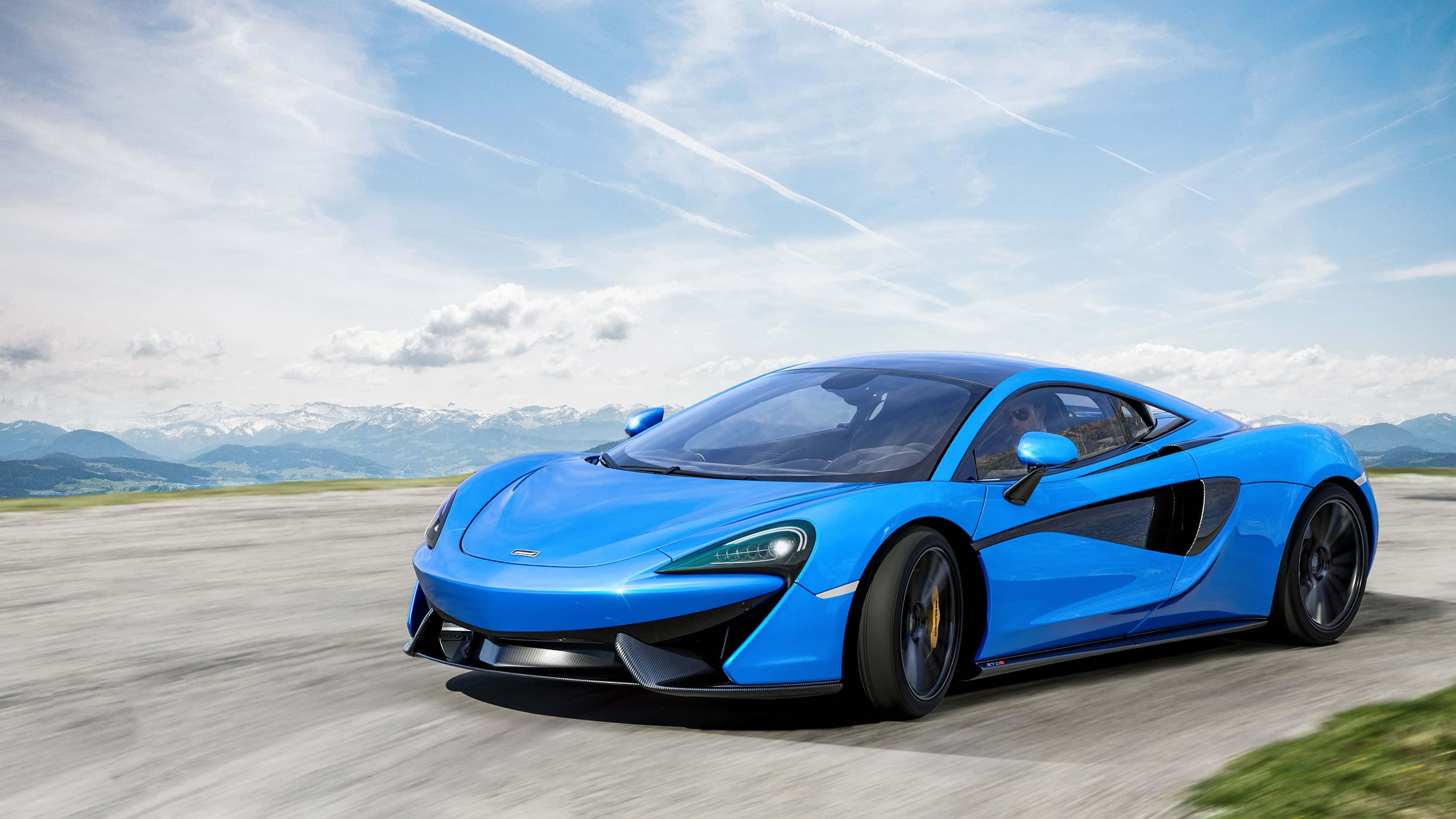 麥拉倫 桌布 壁紙 — 一輛充滿活力的藍色McLaren跑車在明亮的天空下沿著山路飛馳，遠處是白雪皚皚的山峰。