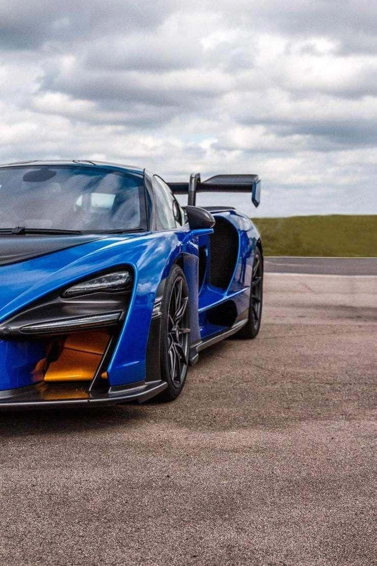 Luxury 桌布 壁紙 — 一輛充滿活力的藍色McLaren Senna超級跑車，帶有橙色點綴，停在灰色瀝青賽道上，天空多雲。