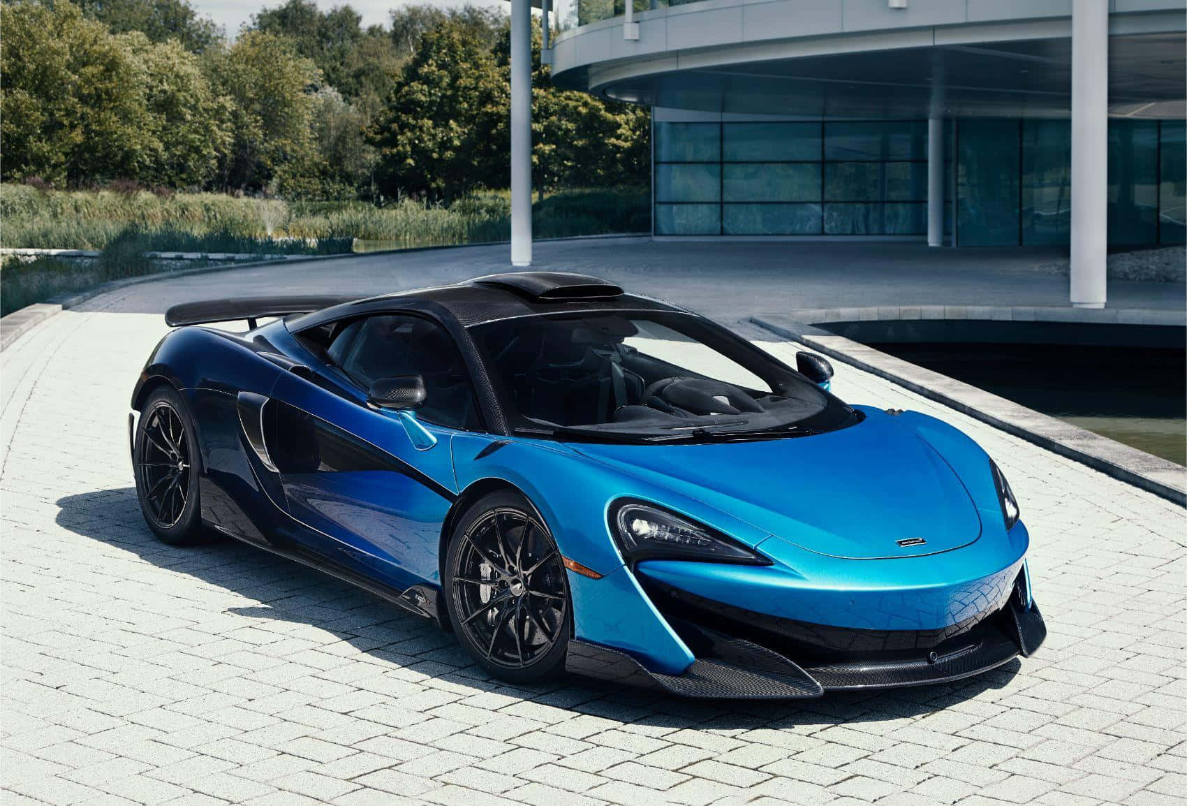 酷車 桌布 壁紙 — 一輛充滿活力的藍色McLaren跑車在明亮的陽光下閃閃發光，停放在現代玻璃建築旁的鋪砌車道上。