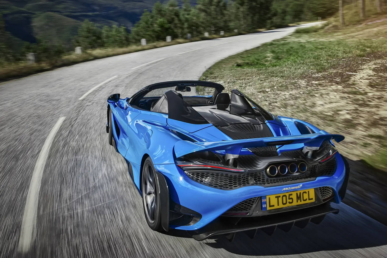 跑車 桌布 壁紙 — 一輛充滿活力的藍色McLaren 570S Spider在蜿蜒的山路上疾馳，帶有模糊的動感和茂密的綠意。