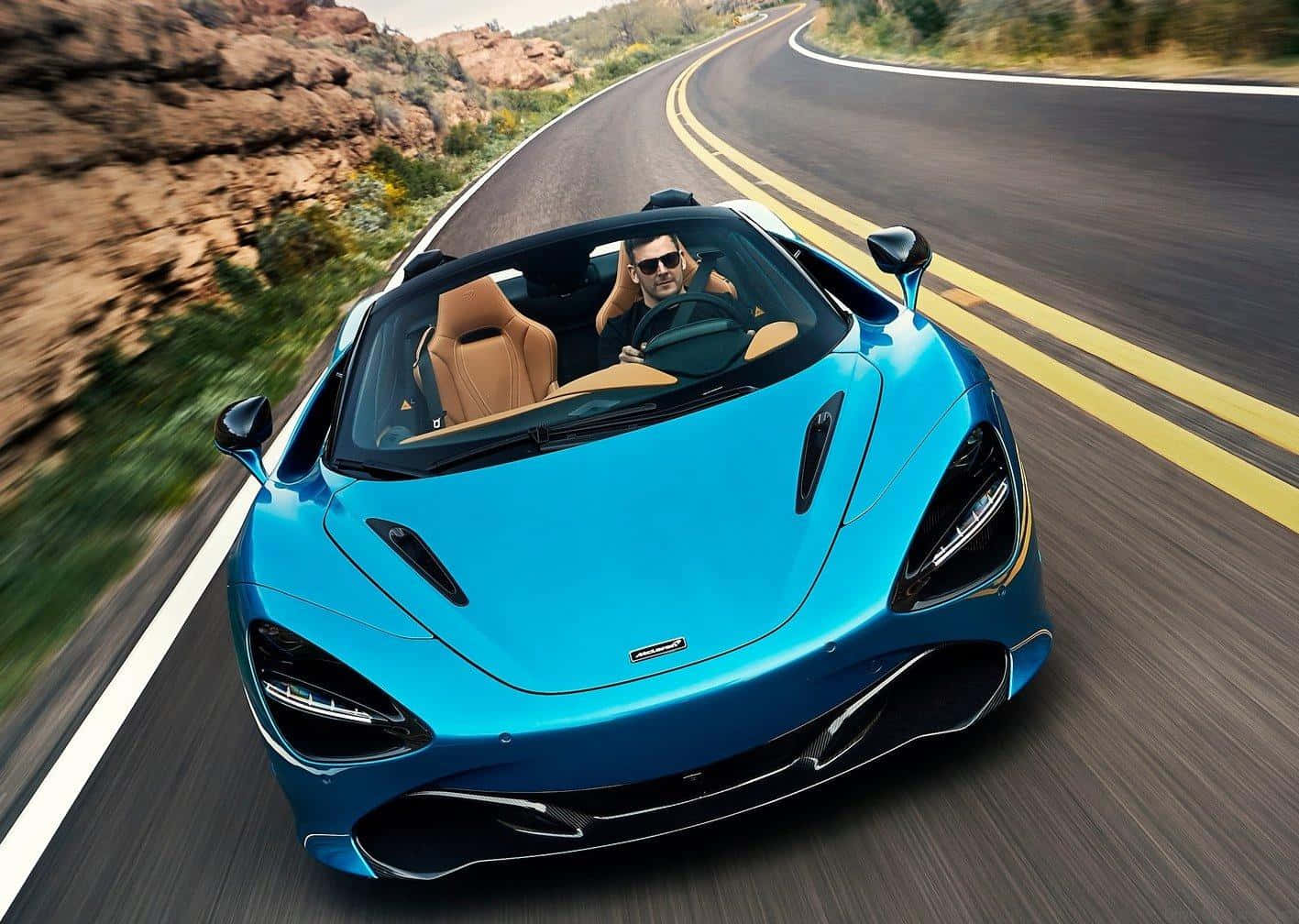 山景 桌布 壁紙 — 一輛充滿活力的藍色McLaren跑車在蜿蜒的沙漠公路上疾馳，司機戴著太陽眼鏡享受駕駛樂趣。