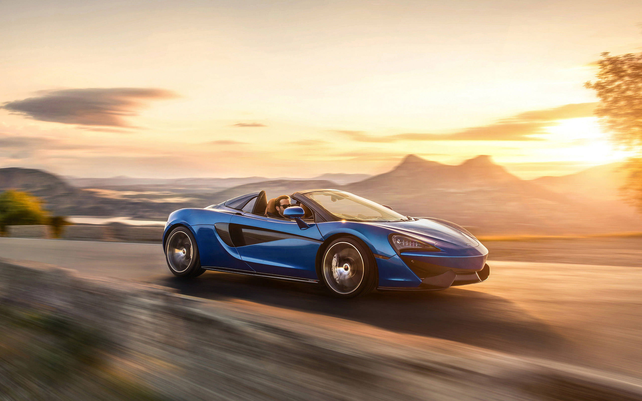 跑車 桌布 壁紙 — 一輛充滿活力的藍色McLaren跑車在日落時分沿著蜿蜒的山路疾馳，帶有模糊的動感和金色光線。
