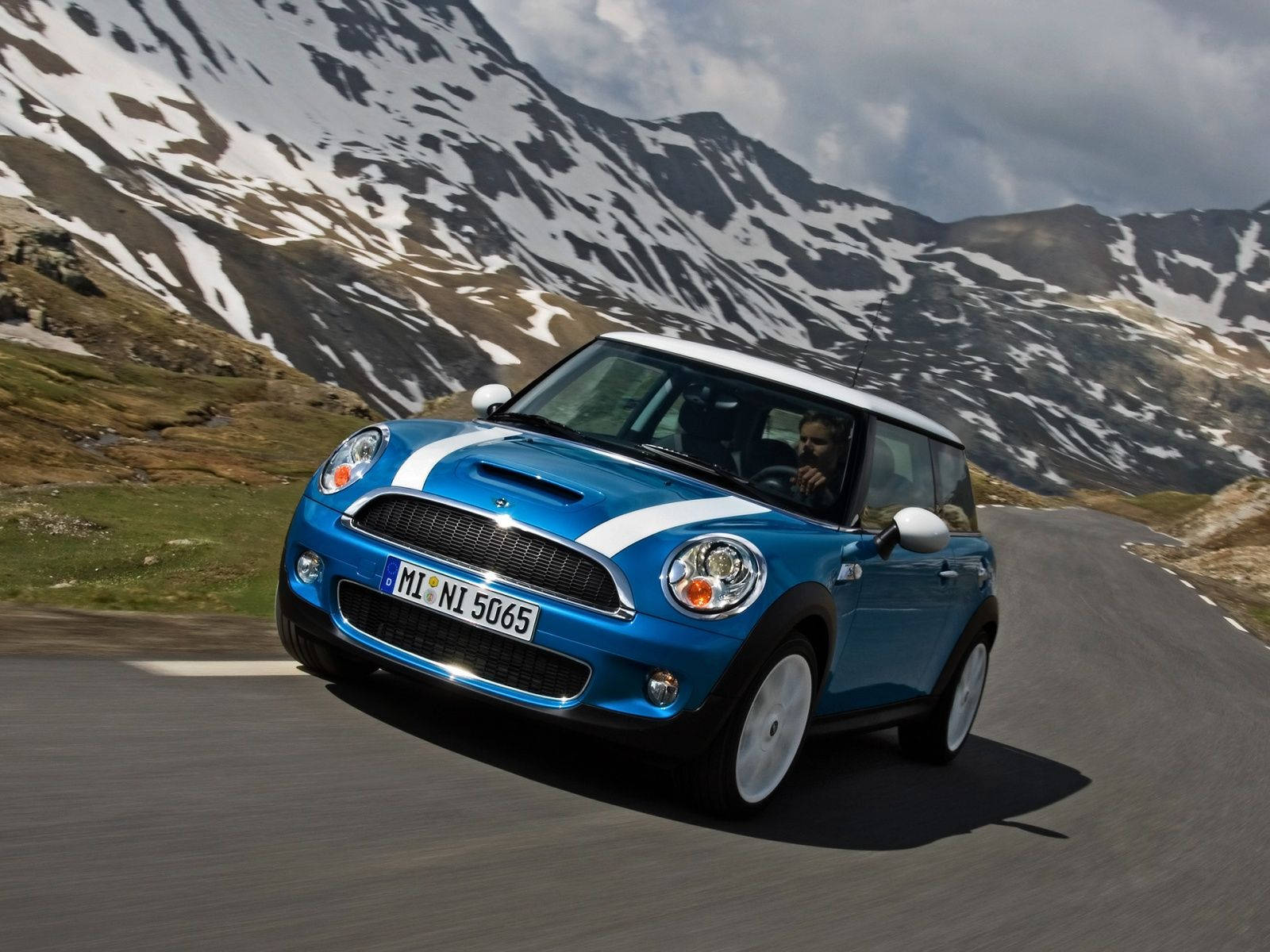 Mini Cooper 桌布 壁紙 — 一輛充滿活力的藍色Mini Cooper，白色車頂和賽車條紋，沿著蜿蜒的山路疾馳，背景是白雪覆蓋的山峰。