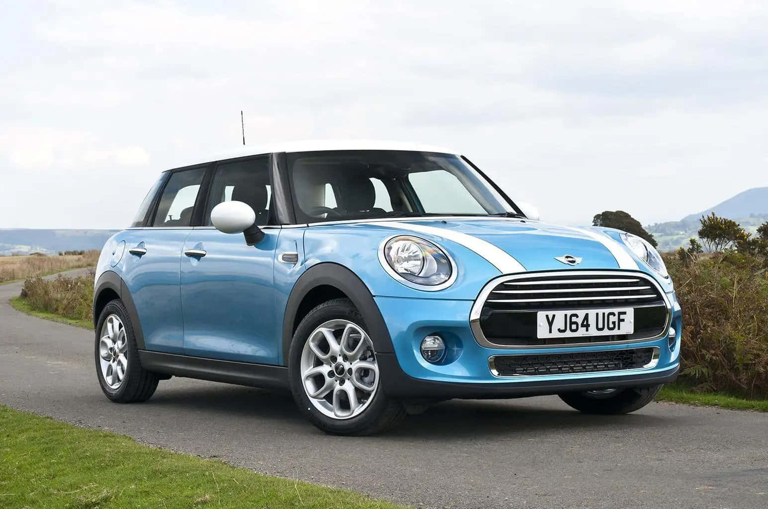 Compact Car 桌布 壁紙 — 一輛充滿活力的藍色Mini Cooper，白色車頂和賽車條紋，停泊在蜿蜒的鄉村道路上，旁邊是草坡，天空多雲。