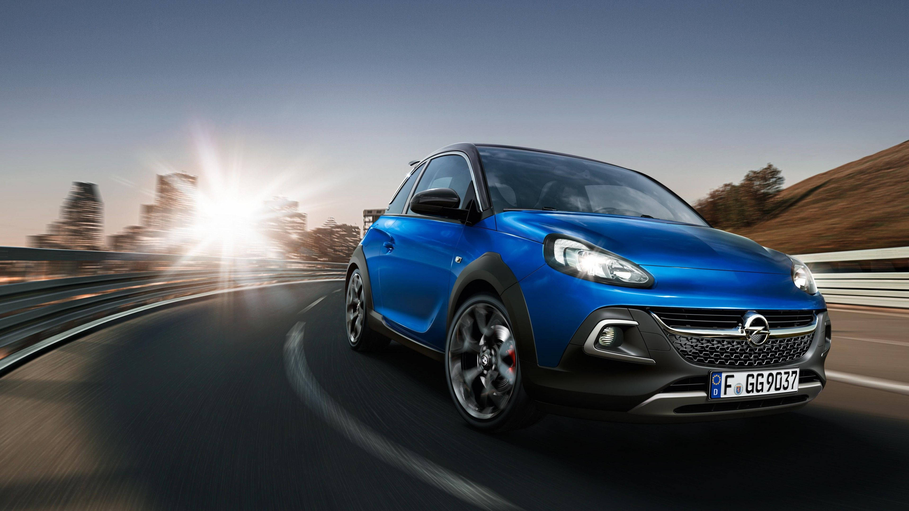 Opel 桌布 壁紙 — 一輛充滿活力的藍色Opel Adam Rocks在蜿蜒的道路上疾馳，背後是模糊的城市景觀和太陽耀斑。