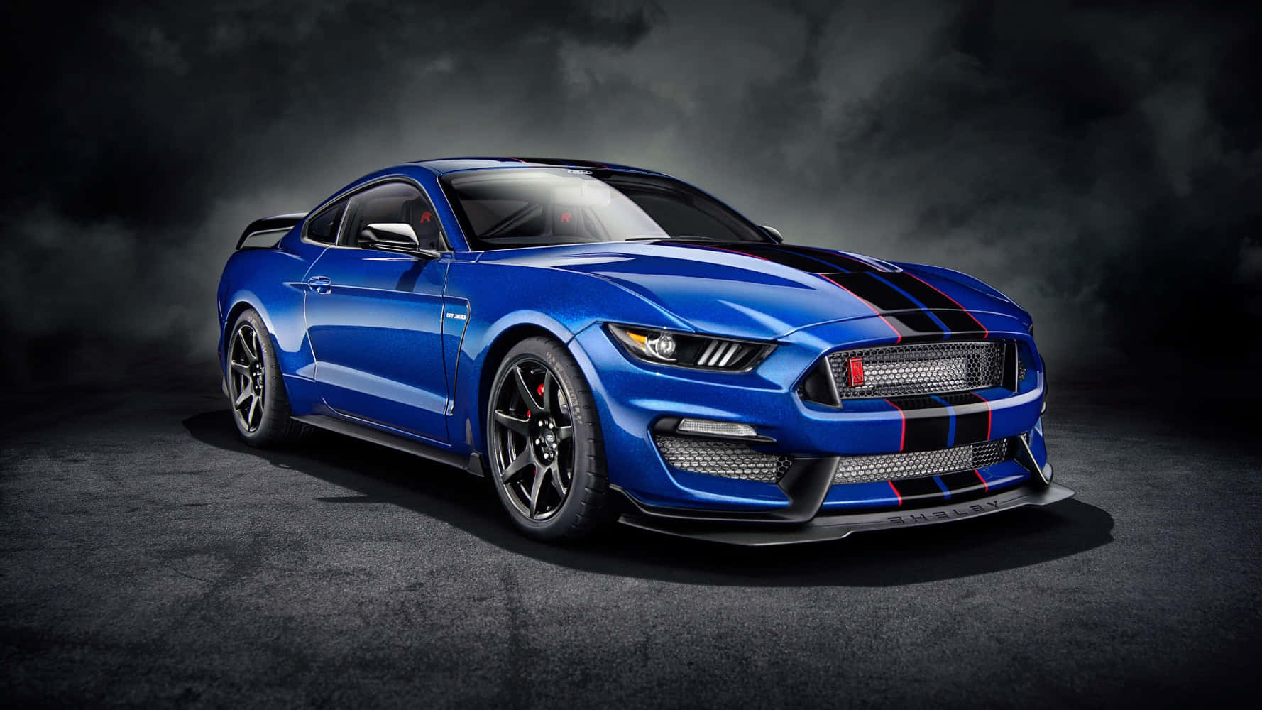 野馬GIF 桌布 壁紙 — 一輛充滿活力的藍色Shelby Mustang GT500，帶有黑色賽車條紋，在煙霧瀰漫的黑暗背景下，在戲劇性的工作室燈光下閃閃發光。