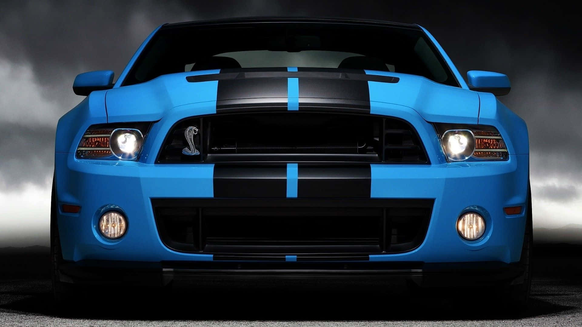 Blue Car 桌布 壁紙 — 一輛充滿活力的藍色Shelby Mustang，帶有黑色賽車條紋，在戲劇性的暴風雨天空下凝視前方。