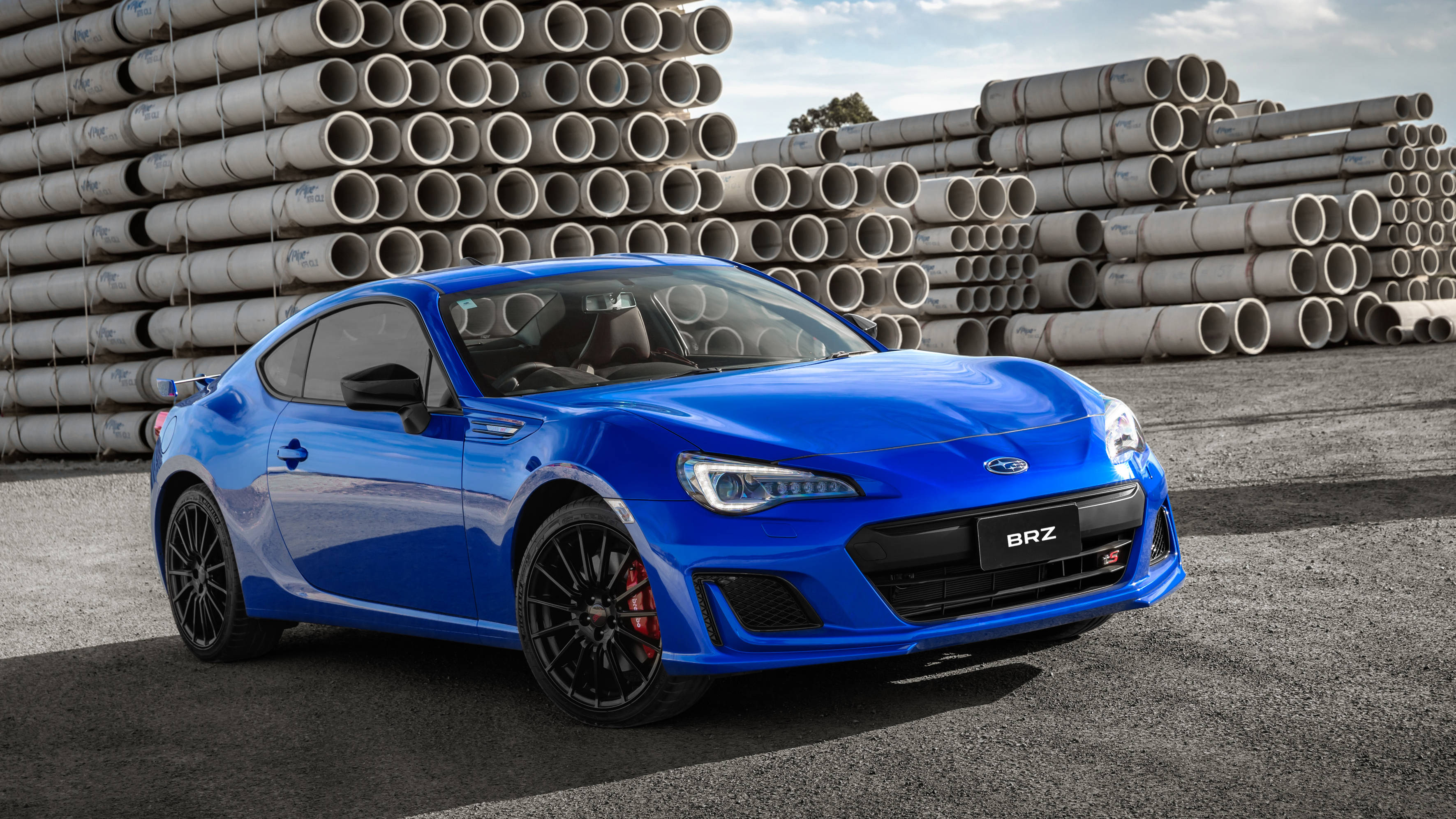 昴 桌布 壁紙 — 一輛充滿活力的藍色Subaru BRZ跑車停在柏油路上，背景是明亮的天空下堆疊的灰色管道。
