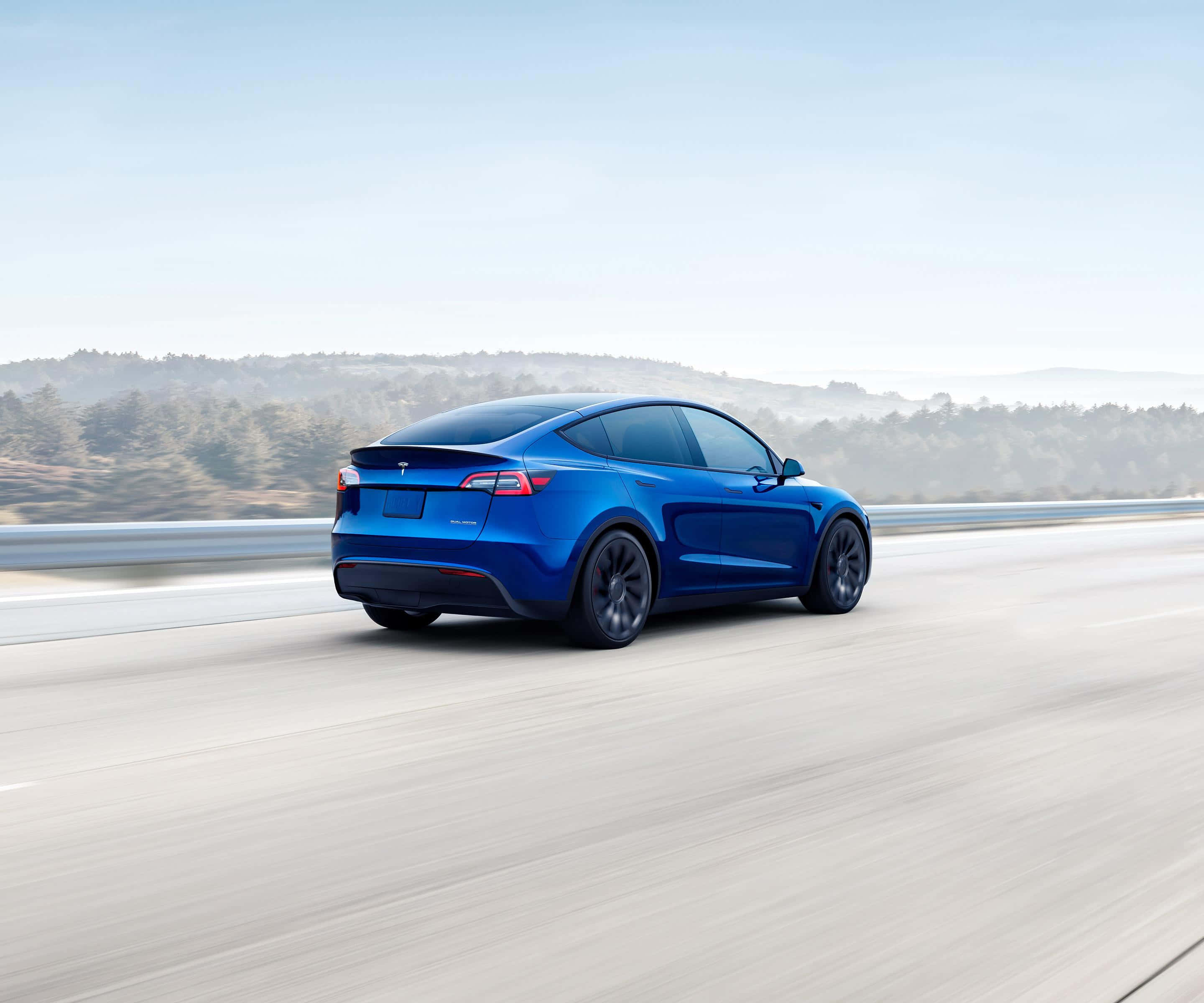 Blue Car 桌布 壁紙 — 一輛充滿活力的藍色Tesla Model Y在高速公路上快速行駛，運動模糊，背景是朦朧的森林山坡和蒼白的天空。