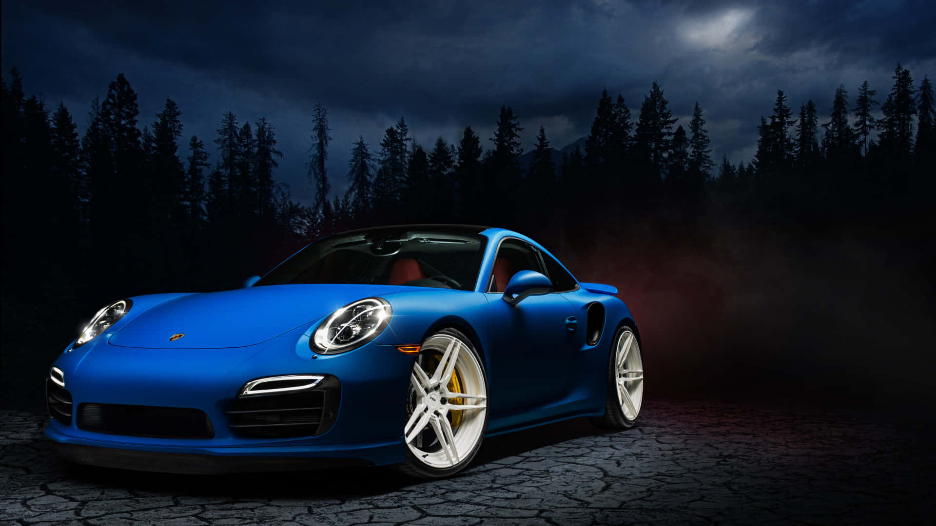 保時捷 桌布 壁紙 — 一輛充滿活力的藍色保時捷911 Turbo S在月光下閃耀，行駛在裂開的道路上，周圍是黑暗的松樹。