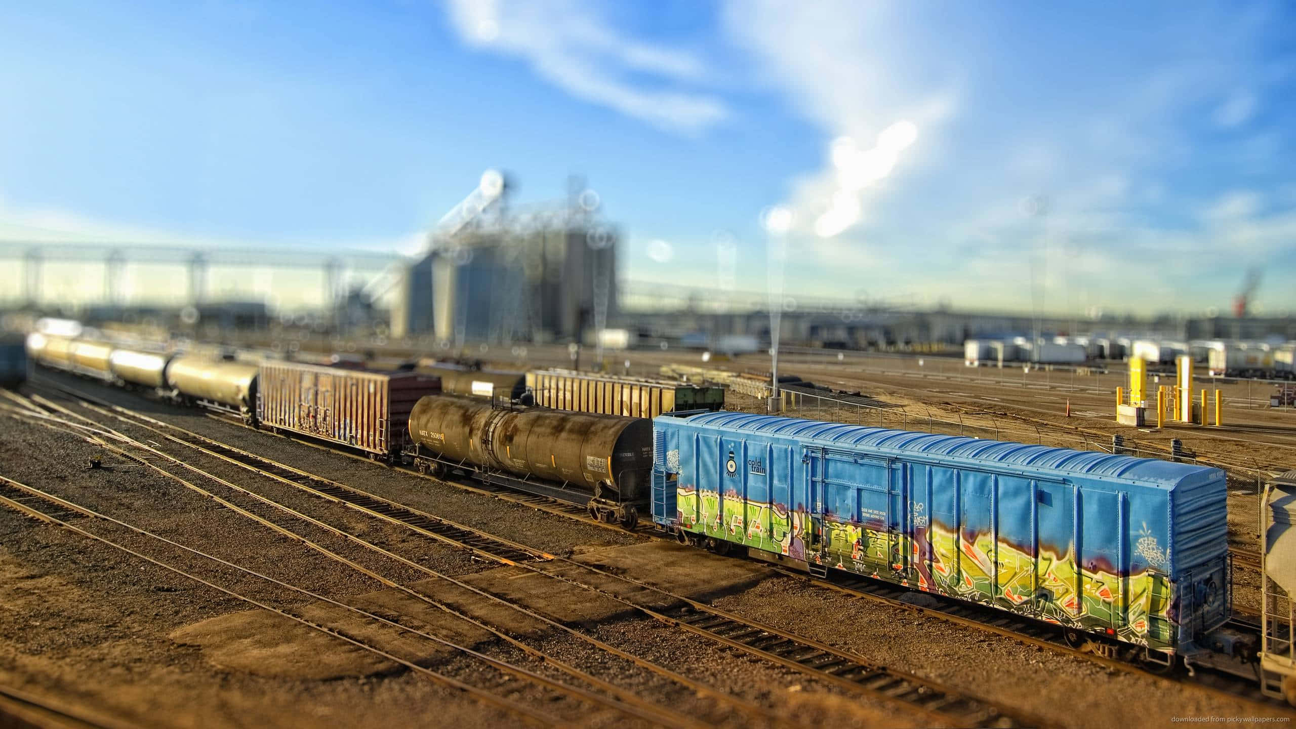 Rust 桌布 壁紙 — 一輛充滿活力的藍色塗鴉火車車廂停在生鏽的軌道上，位於陽光明媚的工業場地，天空清澈湛藍。