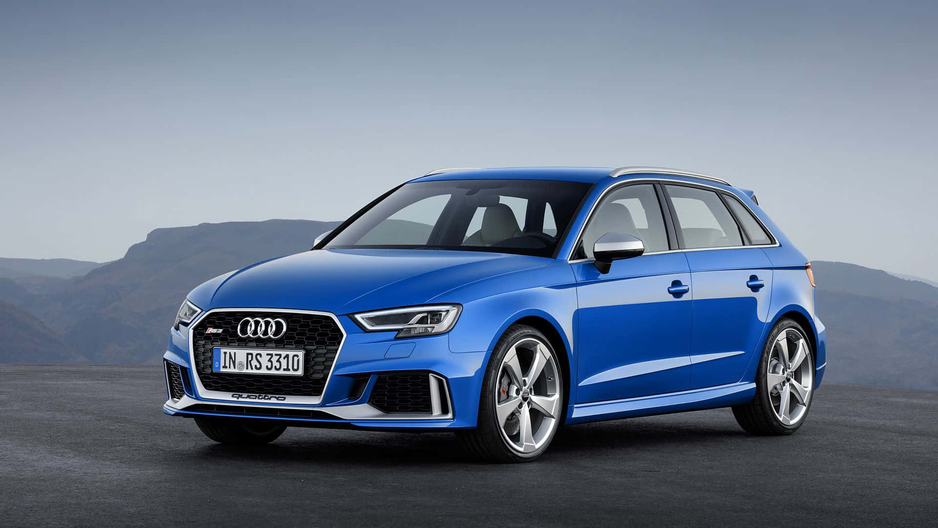 Audi Rs3 桌布 壁紙 — 一輛充滿活力的藍色奧迪RS3 Sportback在日光下閃耀，背景是遠山和晴朗的天空。
