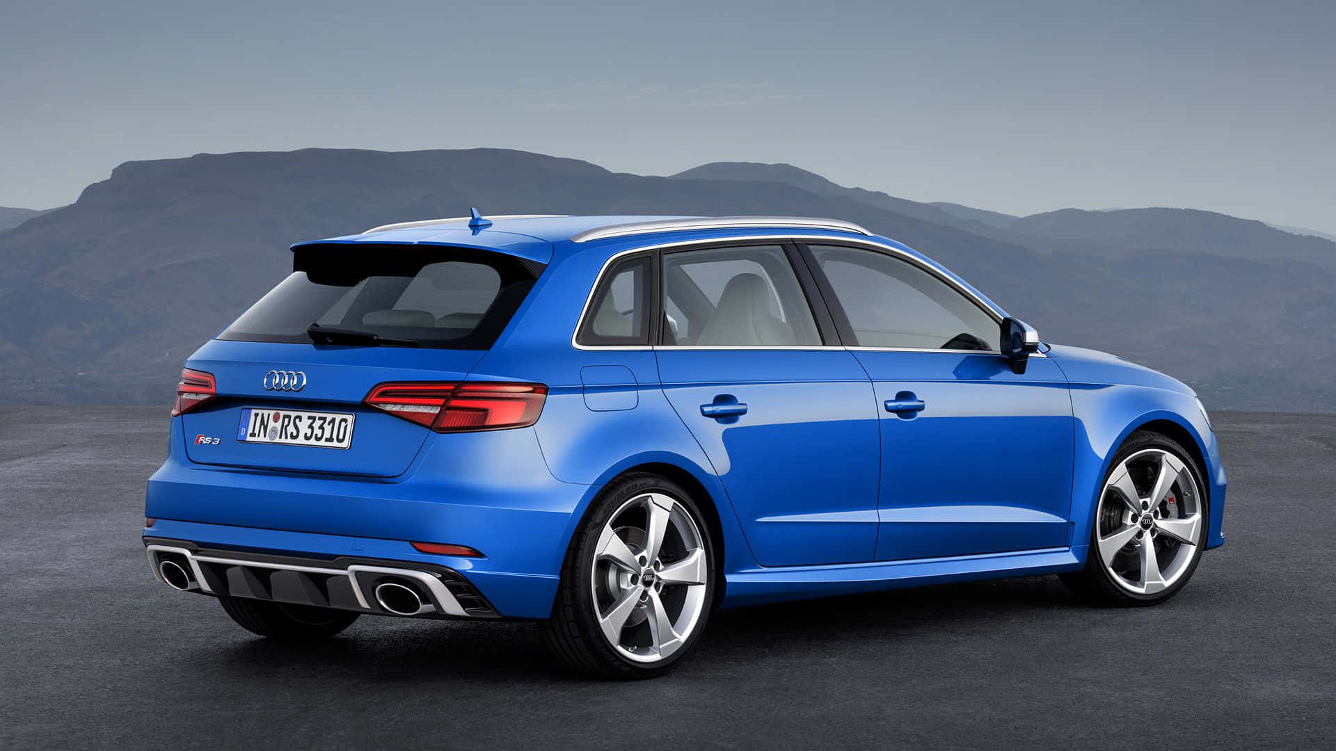 Audi Rs3 桌布 壁紙 — 一輛充滿活力的藍色奧迪RS3 Sportback在日光下閃耀，背景是遠山和晴朗的天空。