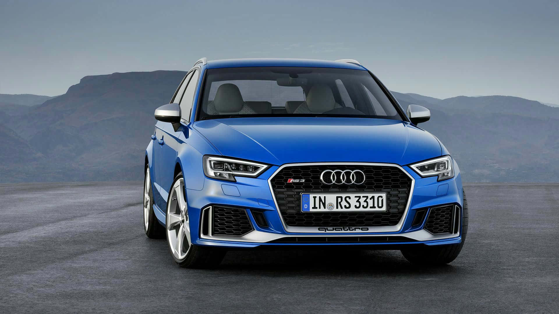 Audi Rs3 桌布 壁紙 — 一輛充滿活力的藍色奧迪RS3跑車停在鋪砌的路面上，遠處山巒在晴朗的天空下。