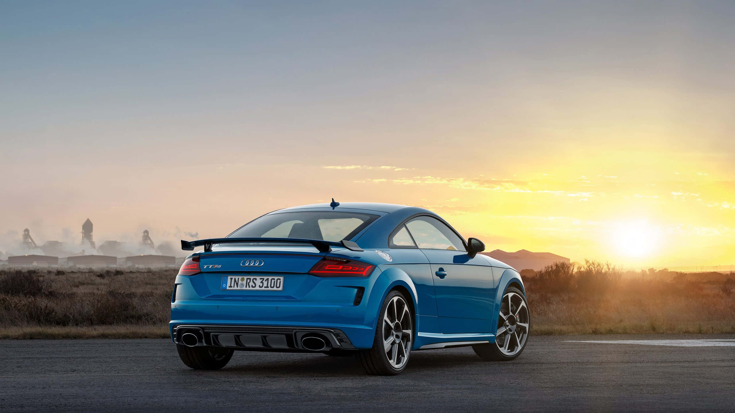 Audi Tt Rs 桌布 壁紙 — 一輛充滿活力的藍色奧迪TT RS雙門跑車停在日落時分的柏油路上，溫暖的金色光線照亮其流線型車尾設計和運動型排氣管。