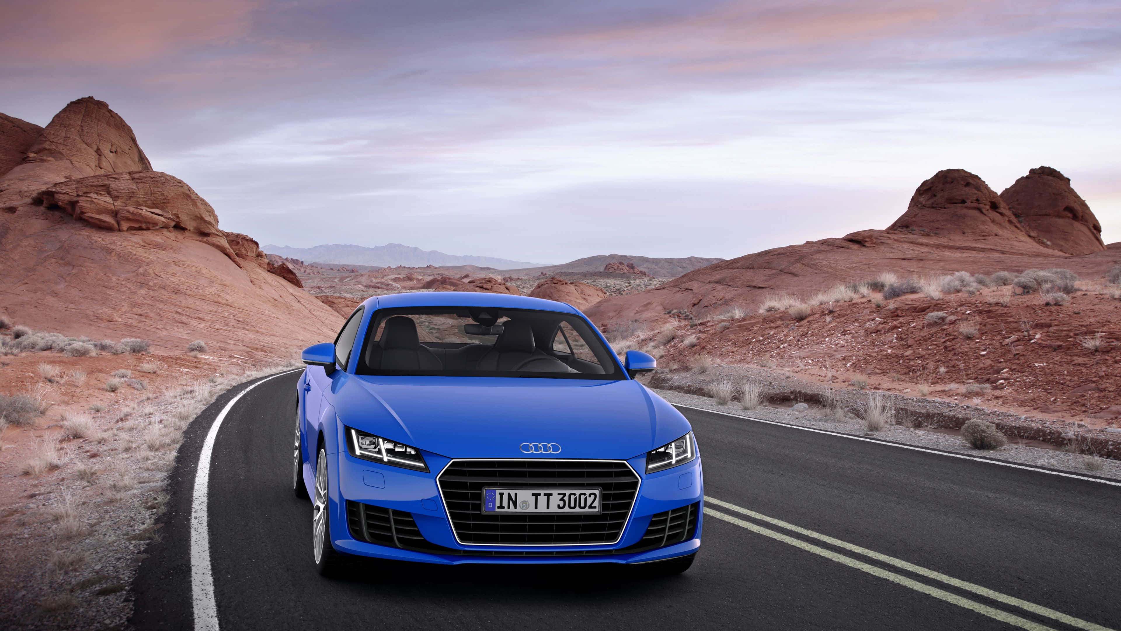 Audi Tt 桌布 壁紙 — 一輛充滿活力的藍色奧迪TT跑車，在柔和的暮色天空下，沿著兩旁有紅色岩層的蜿蜒沙漠公路行駛。