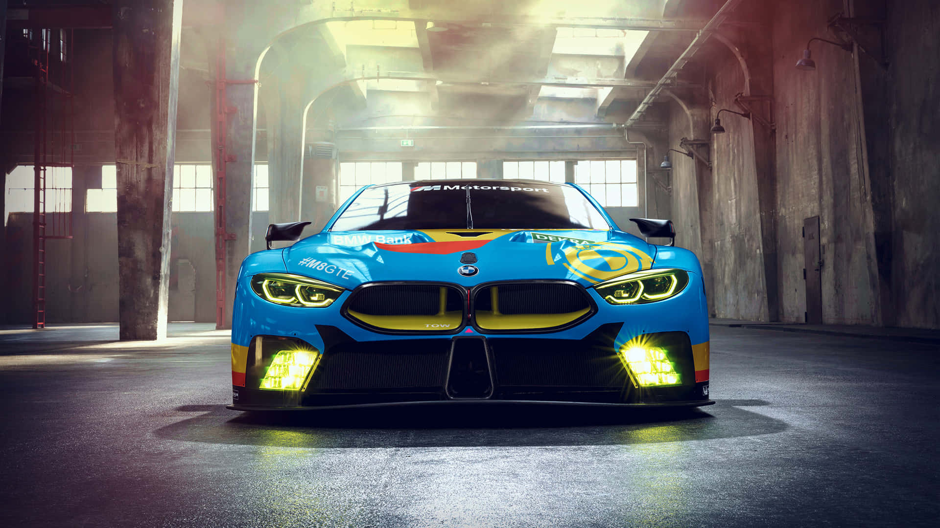 BMW 動圖 桌布 壁紙 — 一輛充滿活力的藍色寶馬賽車，帶有發光的黃色車頭燈，在戲劇性的燈光下主宰著佈滿灰塵的工業倉庫。