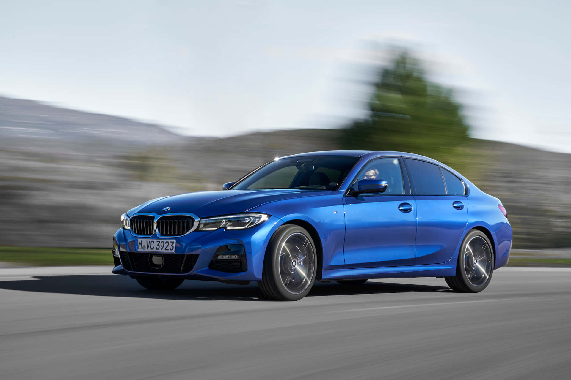 Bmw 3 Series 桌布 壁紙 — 一輛充滿活力的藍色寶馬轎車沿著蜿蜒的道路疾馳，背景是模糊的綠色山丘和天空。