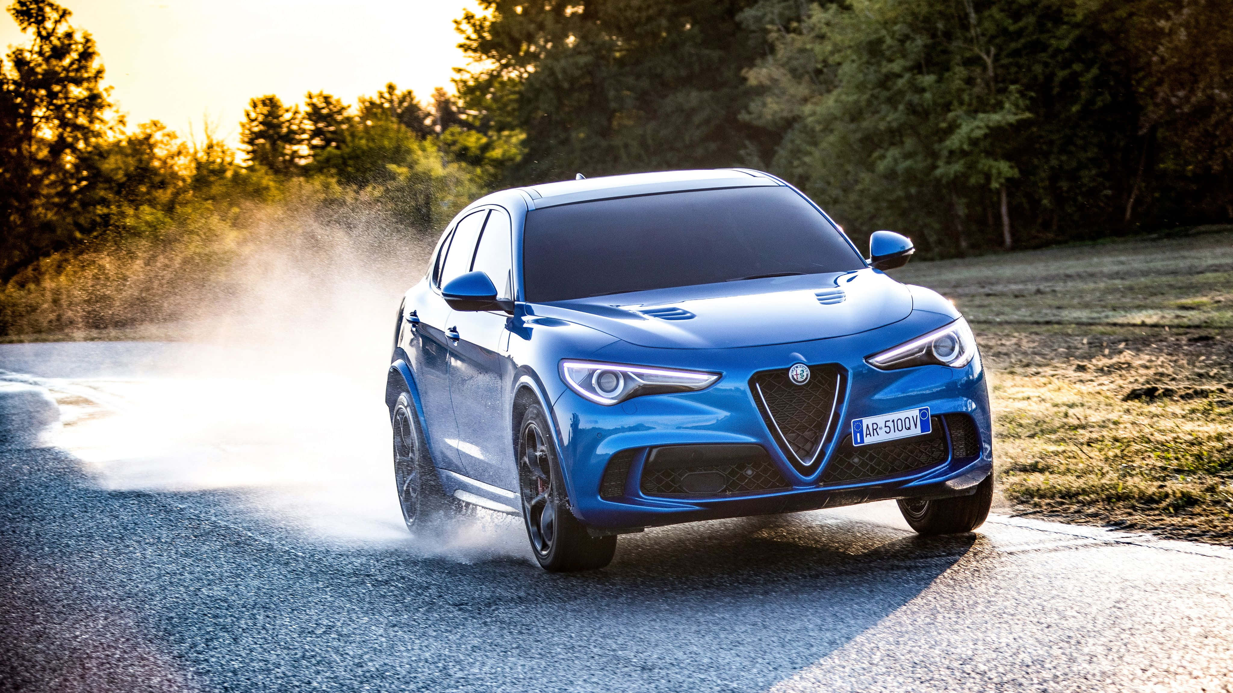 Alfa Romeo Stelvio 桌布 壁紙 — 一輛充滿活力的藍色愛快羅密歐Stelvio SUV在日落時分駛過濕潤的森林道路，激起水花。