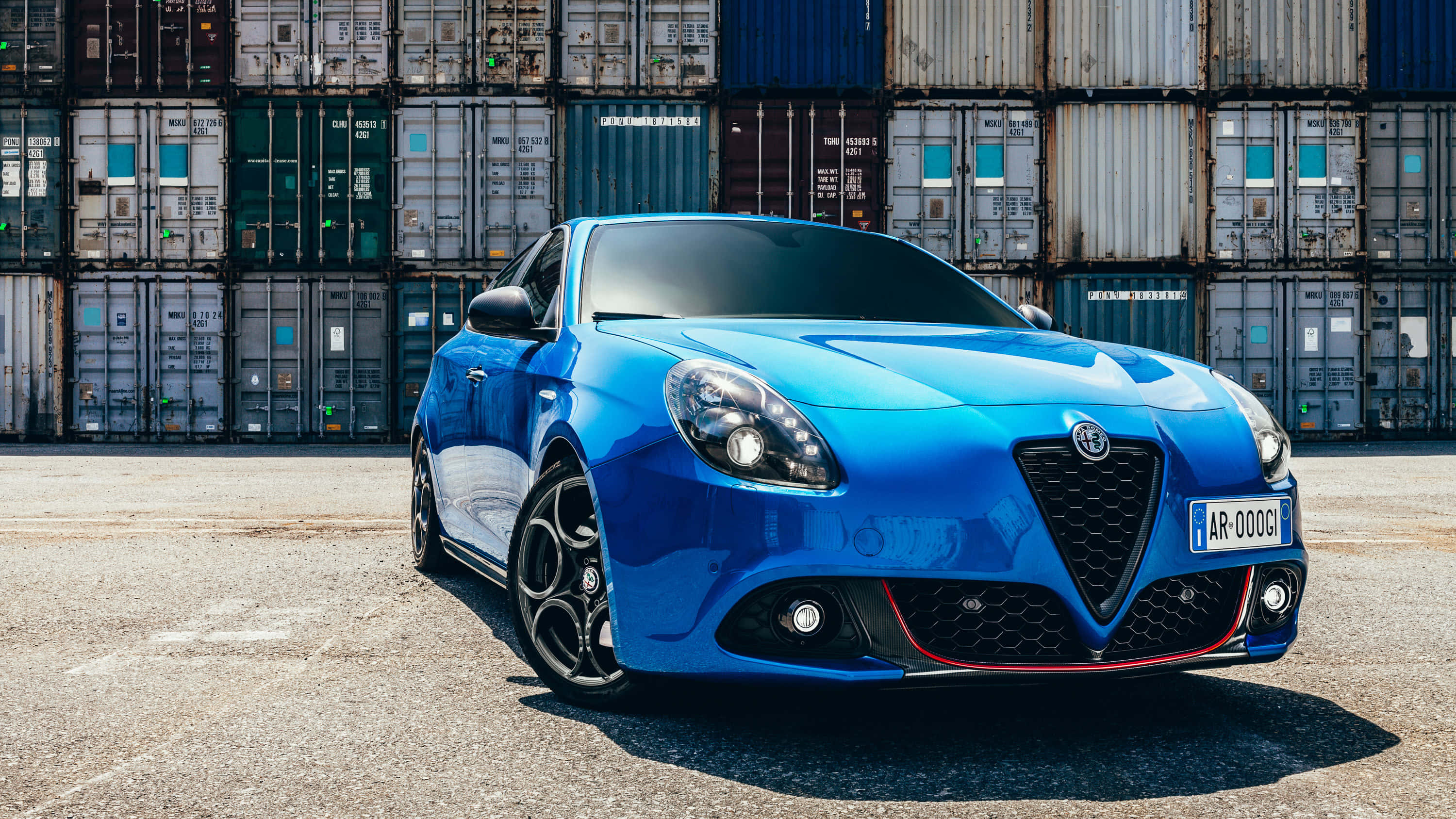 Alfa Romeo Giulietta 桌布 壁紙 — 一輛充滿活力的藍色愛快羅密歐跑車在明亮的陽光下閃閃發光，停泊在一堆高聳的、歷經風霜的貨運貨櫃前。