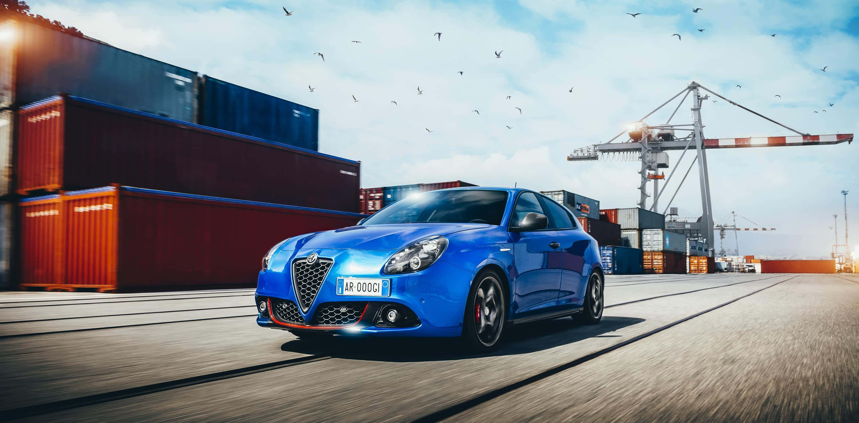 Alfa Romeo Giulietta 桌布 壁紙 — 一輛充滿活力的藍色愛快羅密歐跑車在陽光明媚的港口飛馳，背景是堆疊的貨運貨櫃和起重機。