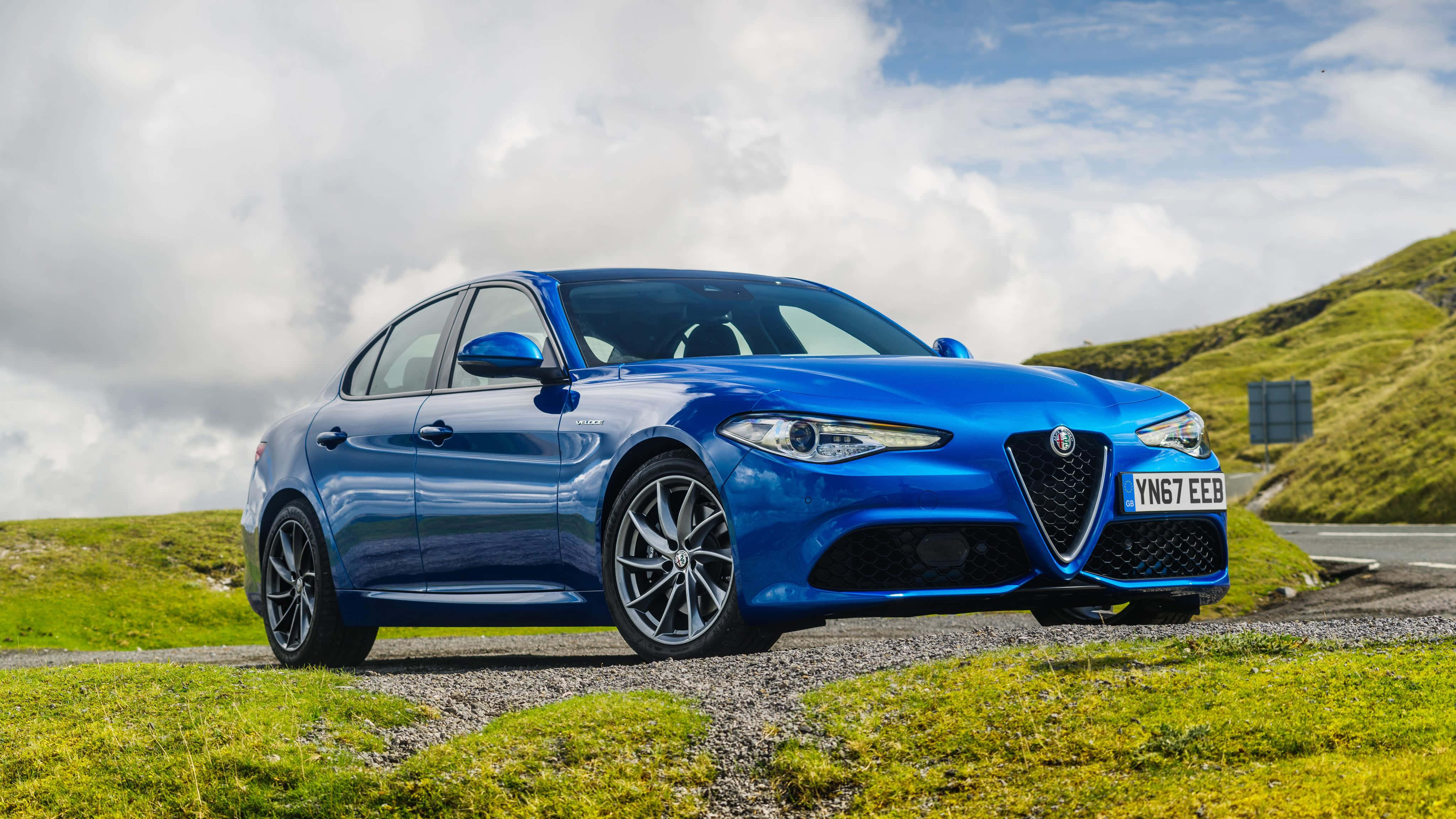 Alfa Romeo Giulia 桌布 壁紙 — 一輛充滿活力的藍色愛快羅密歐轎車停泊在碎石路上，旁邊是草地丘陵，天空多雲。