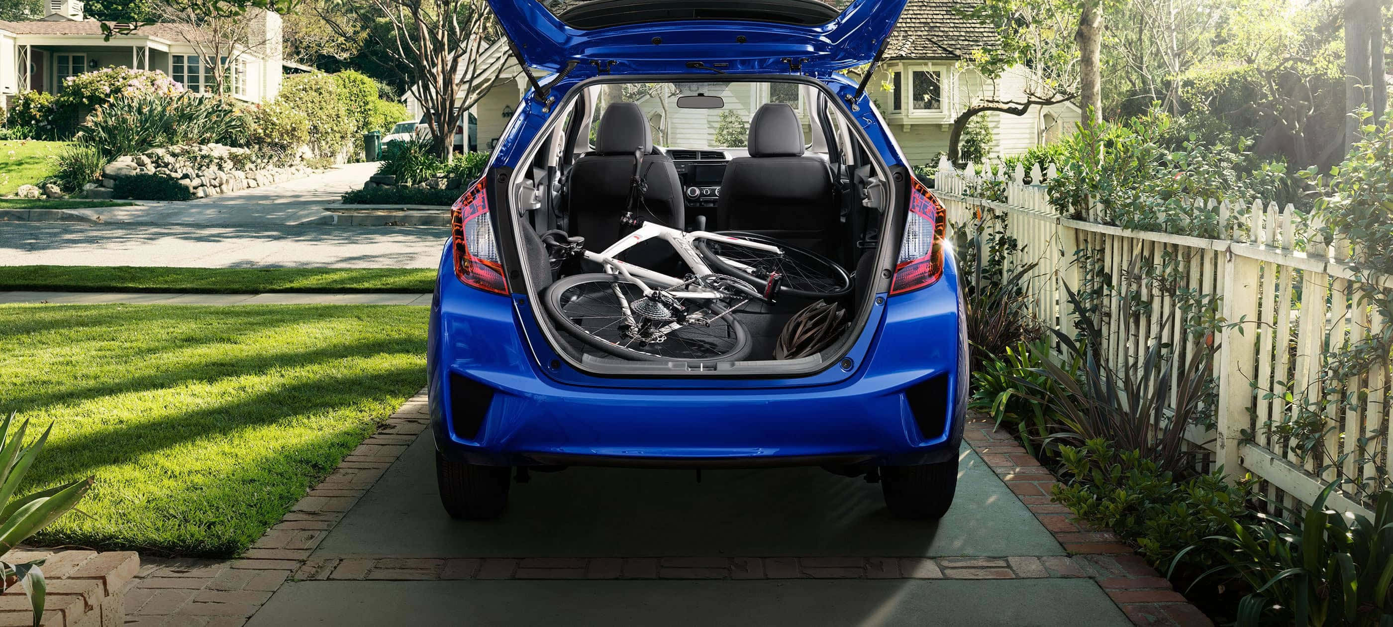 Honda Fit 桌布 壁紙 — 一輛充滿活力的藍色掀背車，尾門打開，露出一輛白色腳踏車停在磚砌車道上，周圍是綠草和白色柵欄。