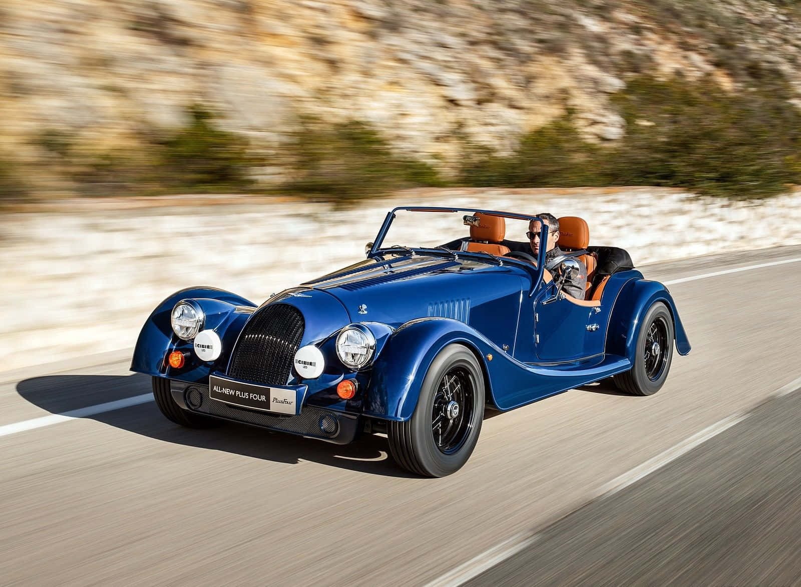 Morgan 桌布 壁紙 — 一輛充滿活力的藍色摩根Plus Four敞篷車在陽光明媚的山路上疾馳，背景是模糊的岩石懸崖。