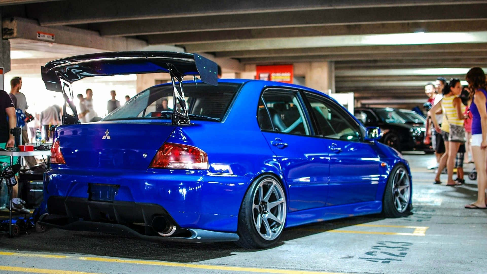 JDM汽車 桌布 壁紙 — 一輛充滿活力的藍色改裝三菱 Lancer Evolution，帶有大型後擾流板，停在混凝土停車場，附近有人。
