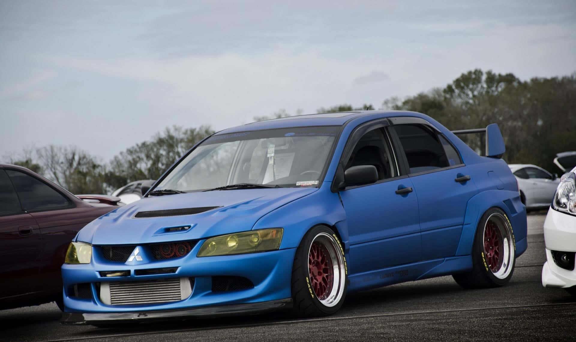 JDM汽車 桌布 壁紙 — 一輛充滿活力的藍色改裝三菱Lancer Evolution，配紅色輪圈，停泊在戶外停車場，天空多雲。
