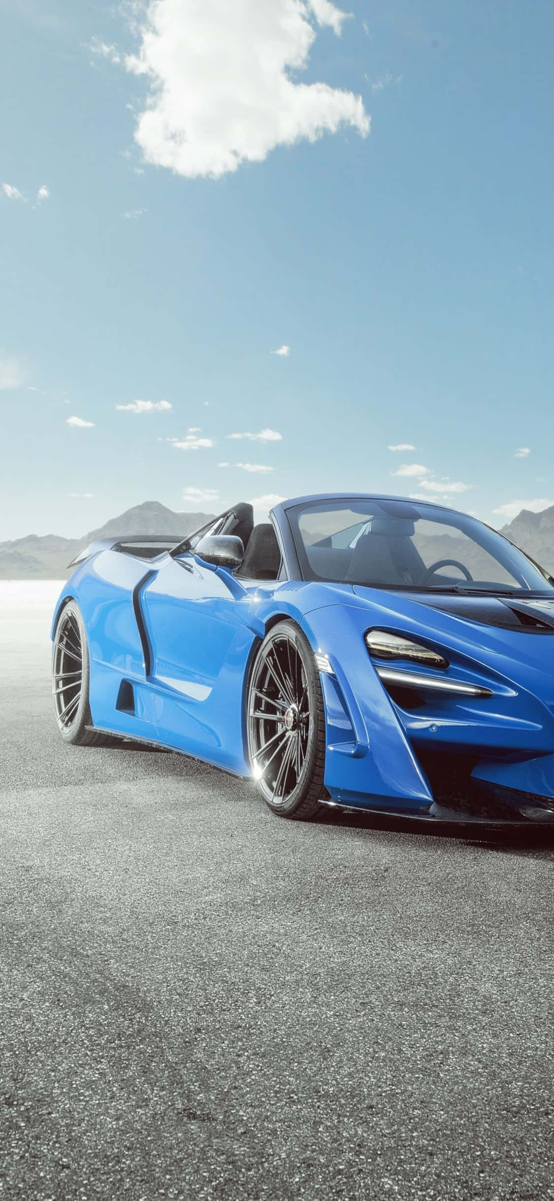 iPhone Xs Mclaren 720s 桌布 壁紙 — 一輛充滿活力的藍色敞篷跑車停放在陽光照耀的柏油路上，遠山在晴朗的天空下。