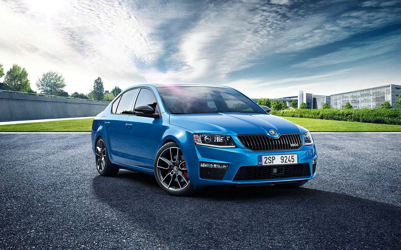 Skoda 桌布 壁紙 — 一輛充滿活力的藍色斯柯達 Octavia RS 轎車在明亮的天空和薄雲下，在鋪砌的道路上閃閃發光，旁邊是綠草和現代建築。