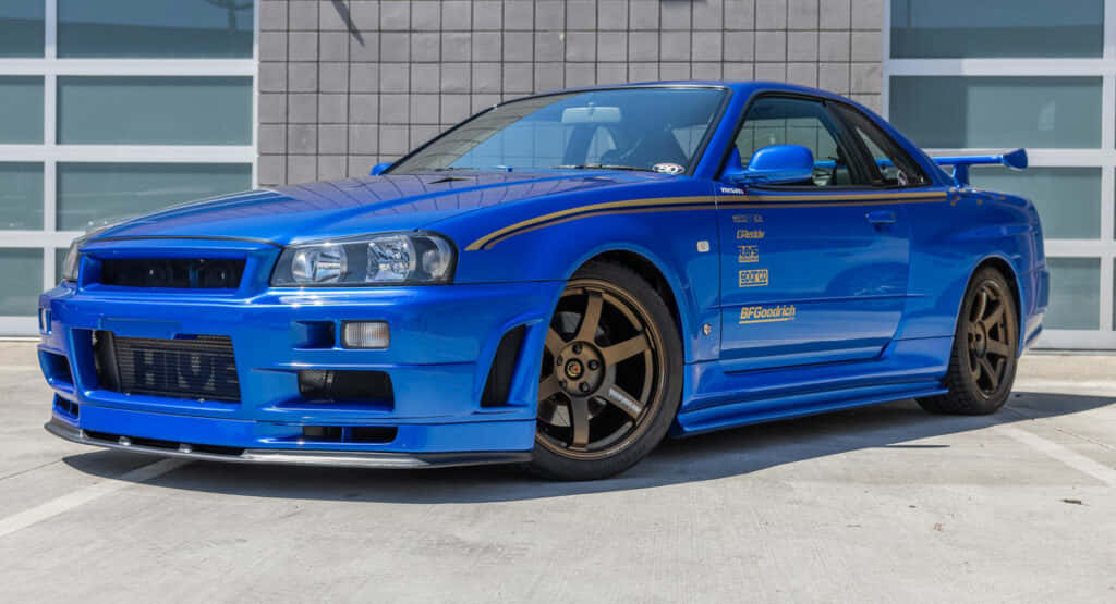 Paul Walker Skyline 桌布 壁紙 — 一輛充滿活力的藍色日產Skyline R34 GT-R跑車停在現代建築前，建築有玻璃窗。