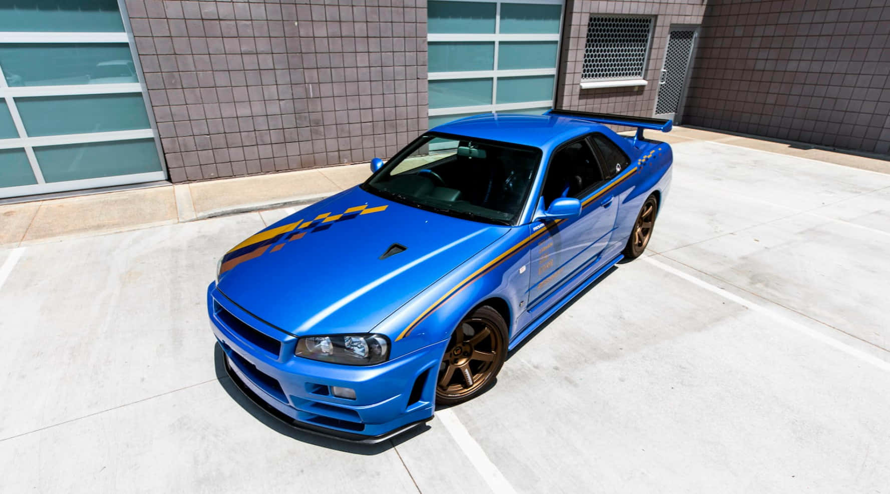 Paul Walker Skyline 桌布 壁紙 — 一輛充滿活力的藍色日產Skyline GT-R，帶有金色賽車條紋，停在明亮的日光下現代混凝土停車場。