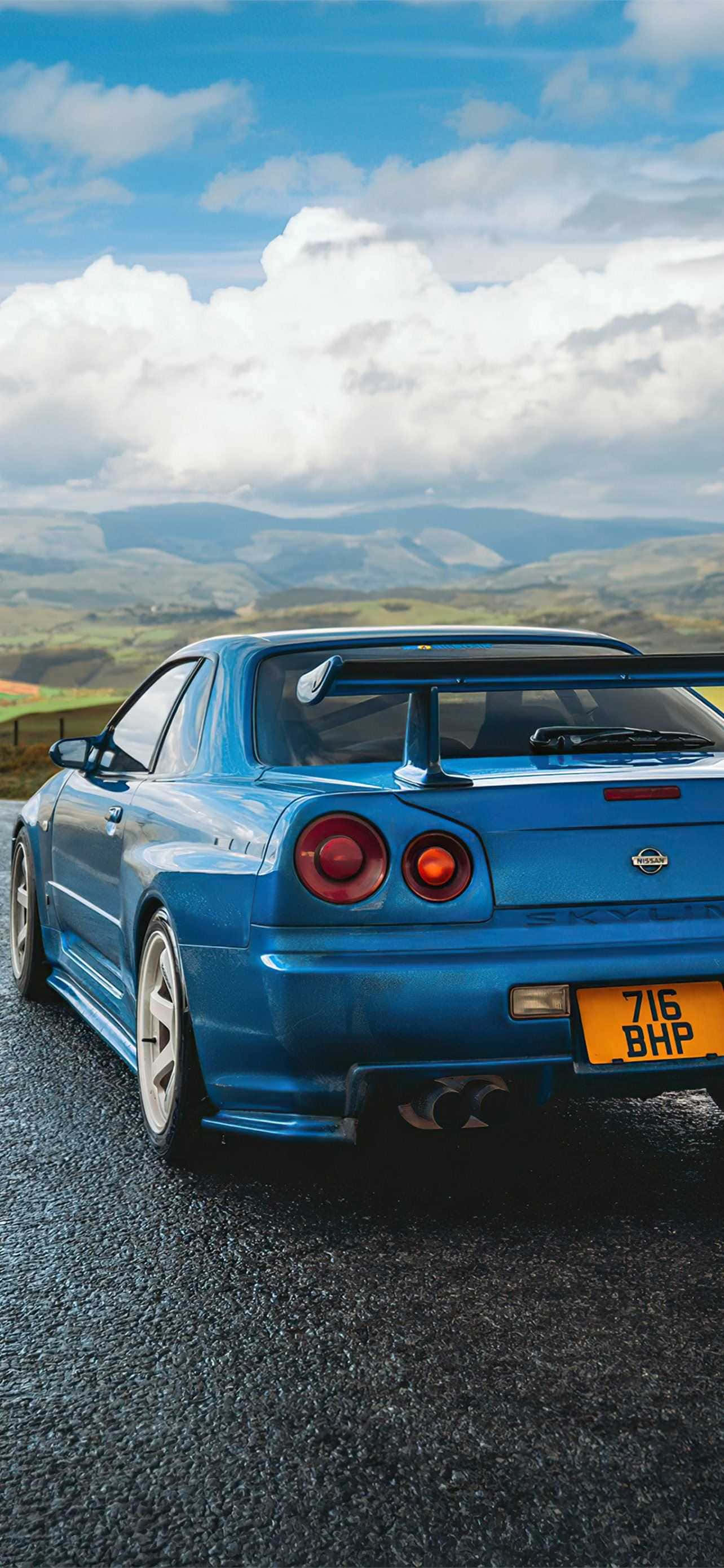 R33 GT-R 桌布 壁紙 — 一輛充滿活力的藍色日產Skyline R34 GT-R停泊在碎石路上，背景是起伏的綠色山丘和蓬鬆的白雲，天空湛藍明亮。