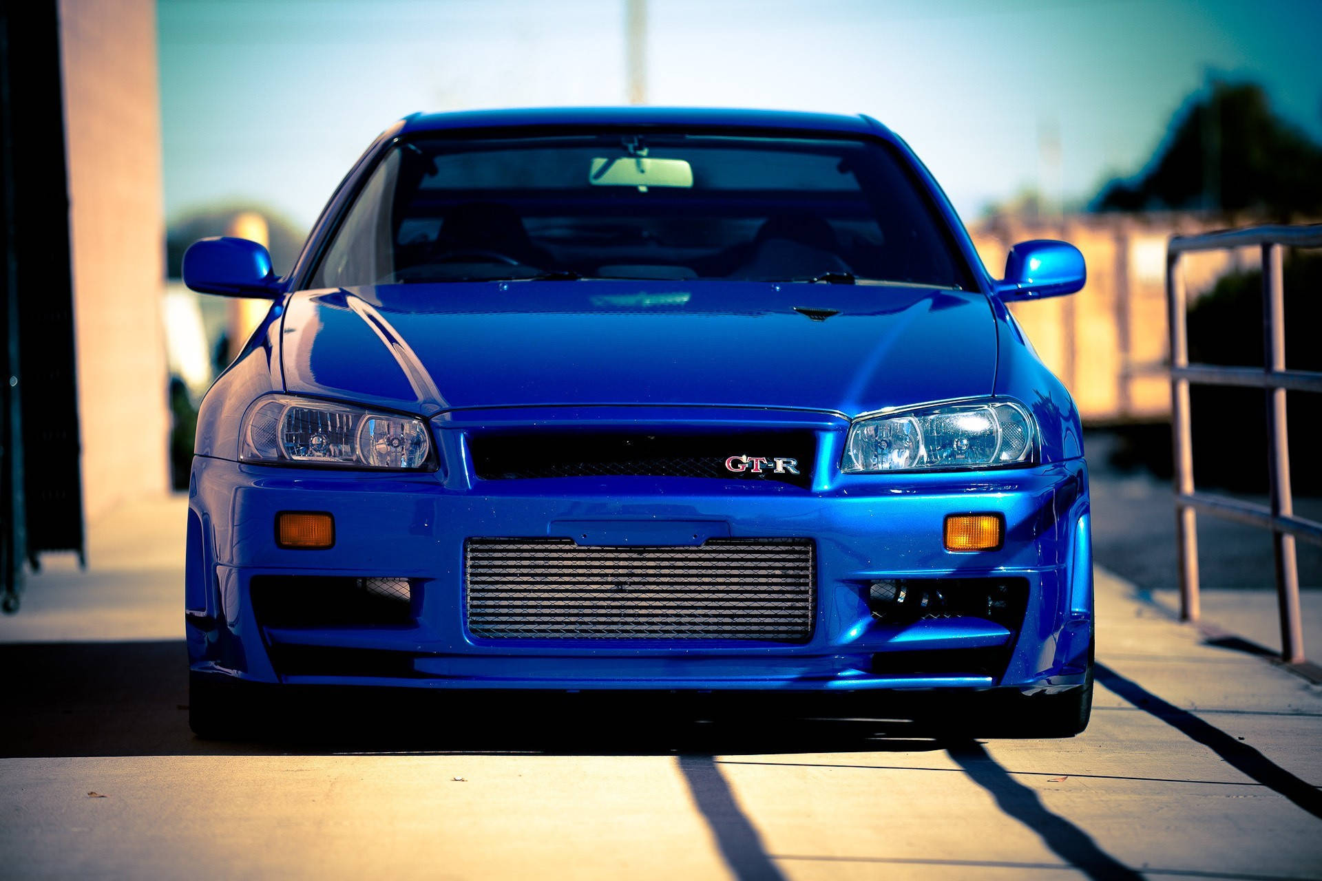 cars 桌布 壁紙 — 一輛充滿活力的藍色日產Skyline GTR停放在陽光普照的混凝土表面上，帶有銳利的陰影和模糊的城市背景。